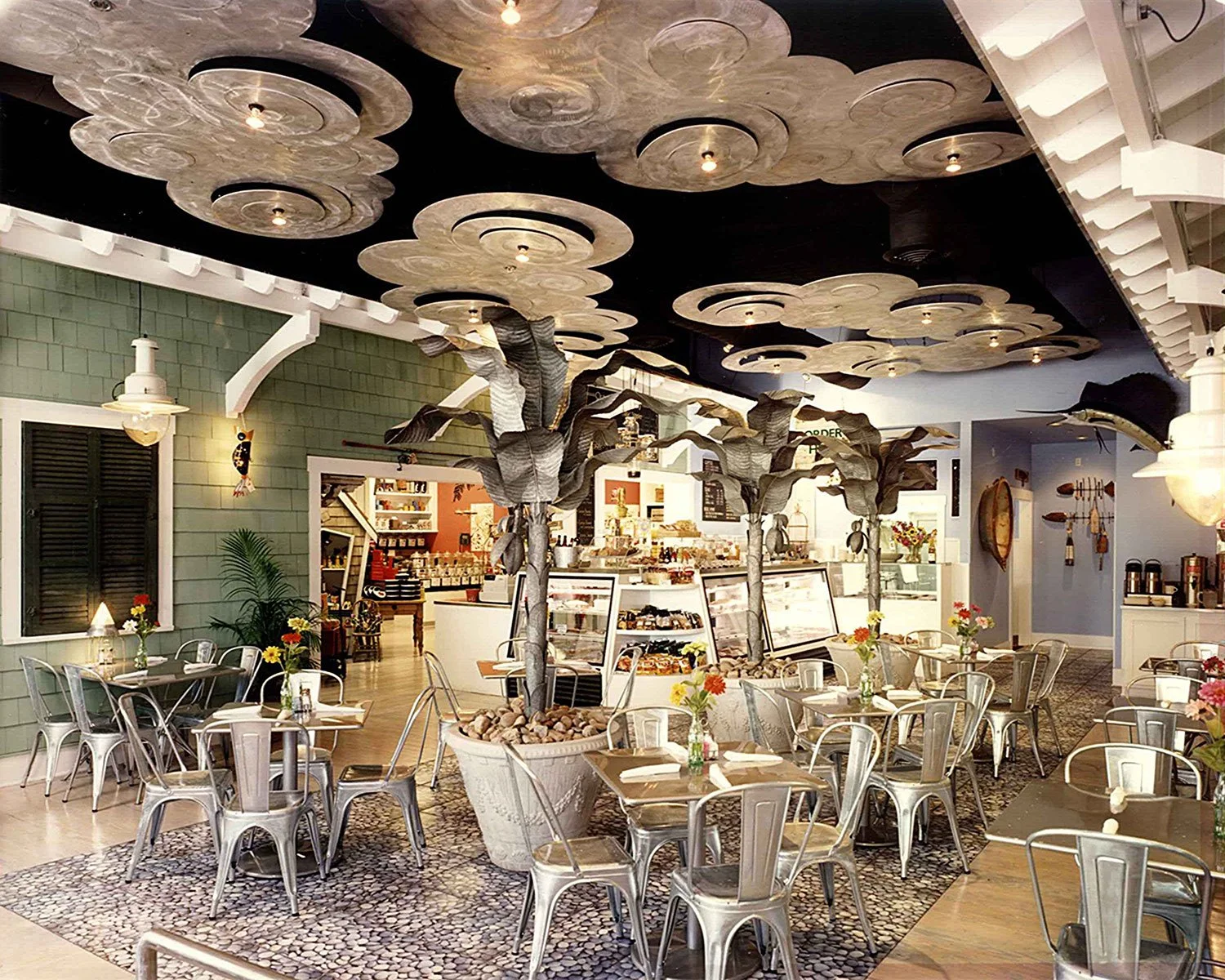 Interior of a cozy cafe or bakery with decorative trees in pots, metallic chairs, and a ceiling with cloud-shaped panels.