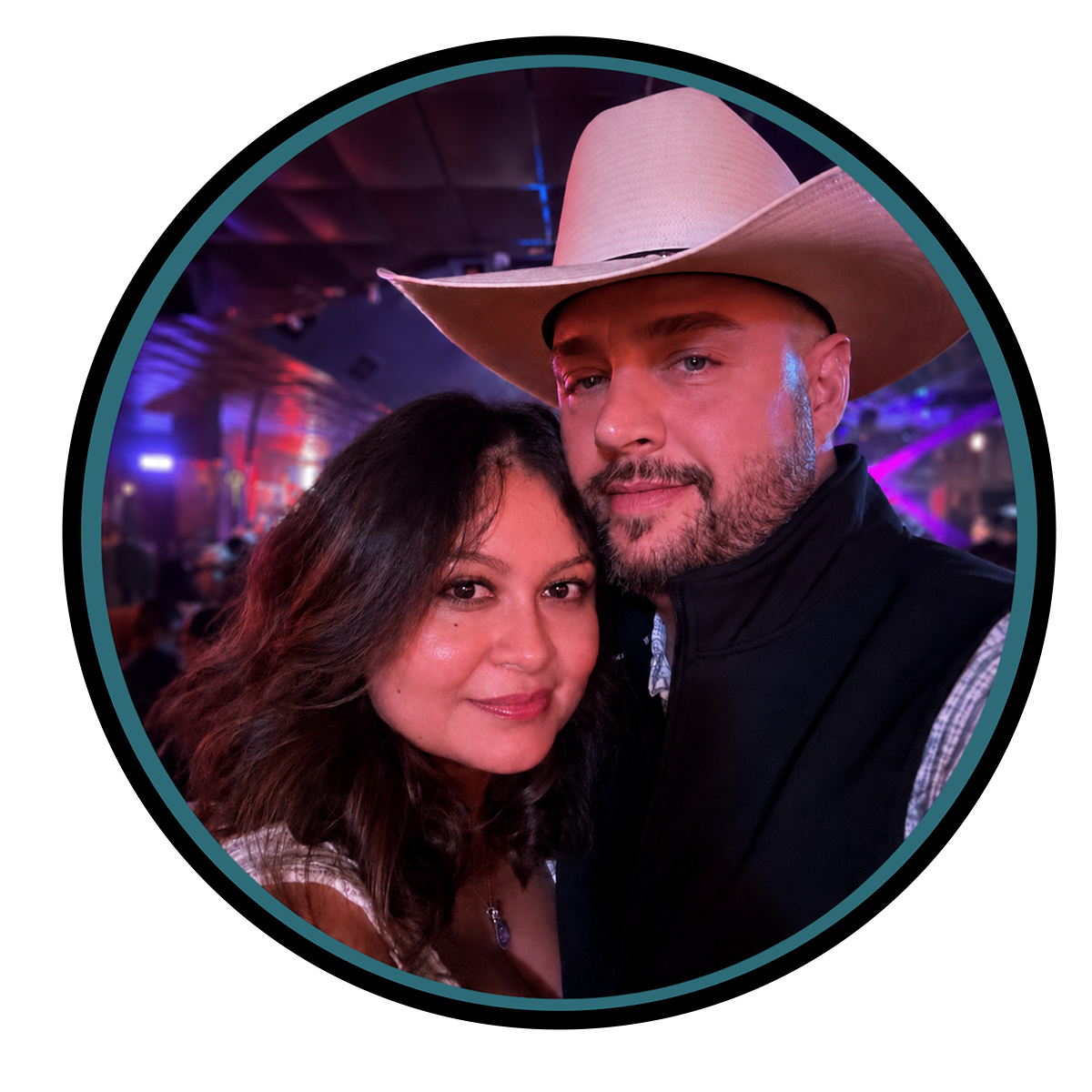 Owners Robert and Alma posing for a photo, with the man wearing a large cowboy hat and the woman having wavy brunette hair, both smiling, with colorful lights in the background.