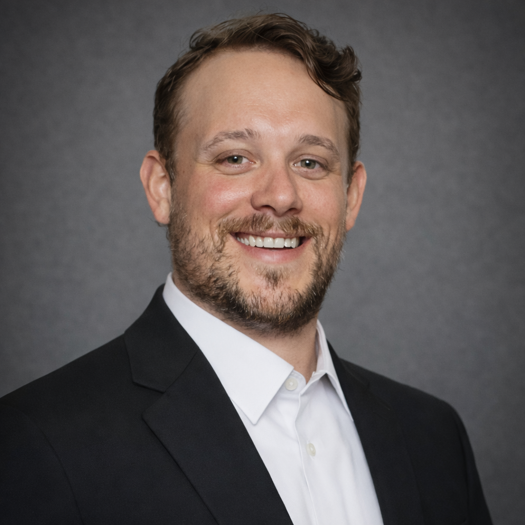 A smiling man with a beard, wearing a black suit and white shirt, against a gray background.