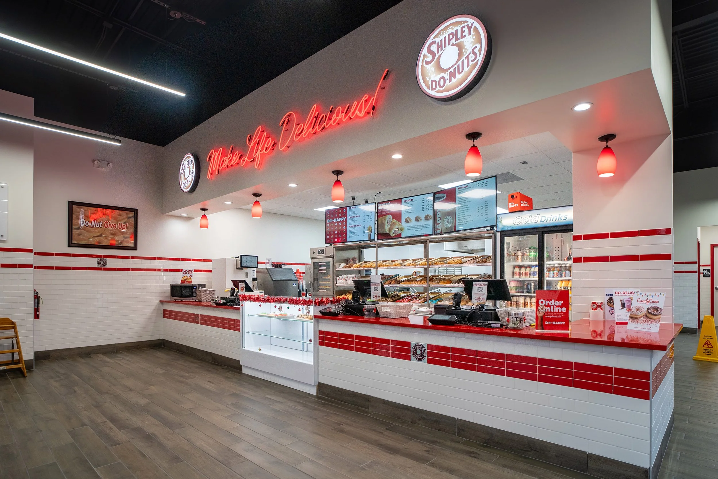Interior of a Shipley's Donuts shop with a red and white color scheme, digital menu screens, donut display case, and neon sign that reads 'Made Life Delicious!'