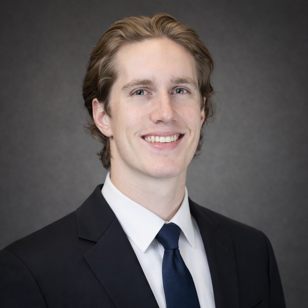 Headshot of a young man with light brown hair wearing a black suit, white shirt, and navy tie, smiling against a dark gray background.