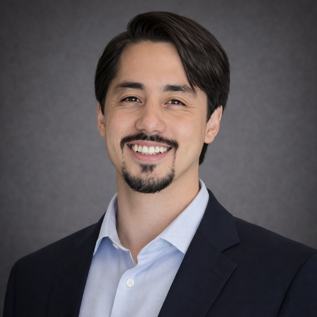 A professional headshot of a smiling man with dark hair, a trimmed beard, and a mustache, wearing a navy blazer and a light blue dress shirt against a gray background.