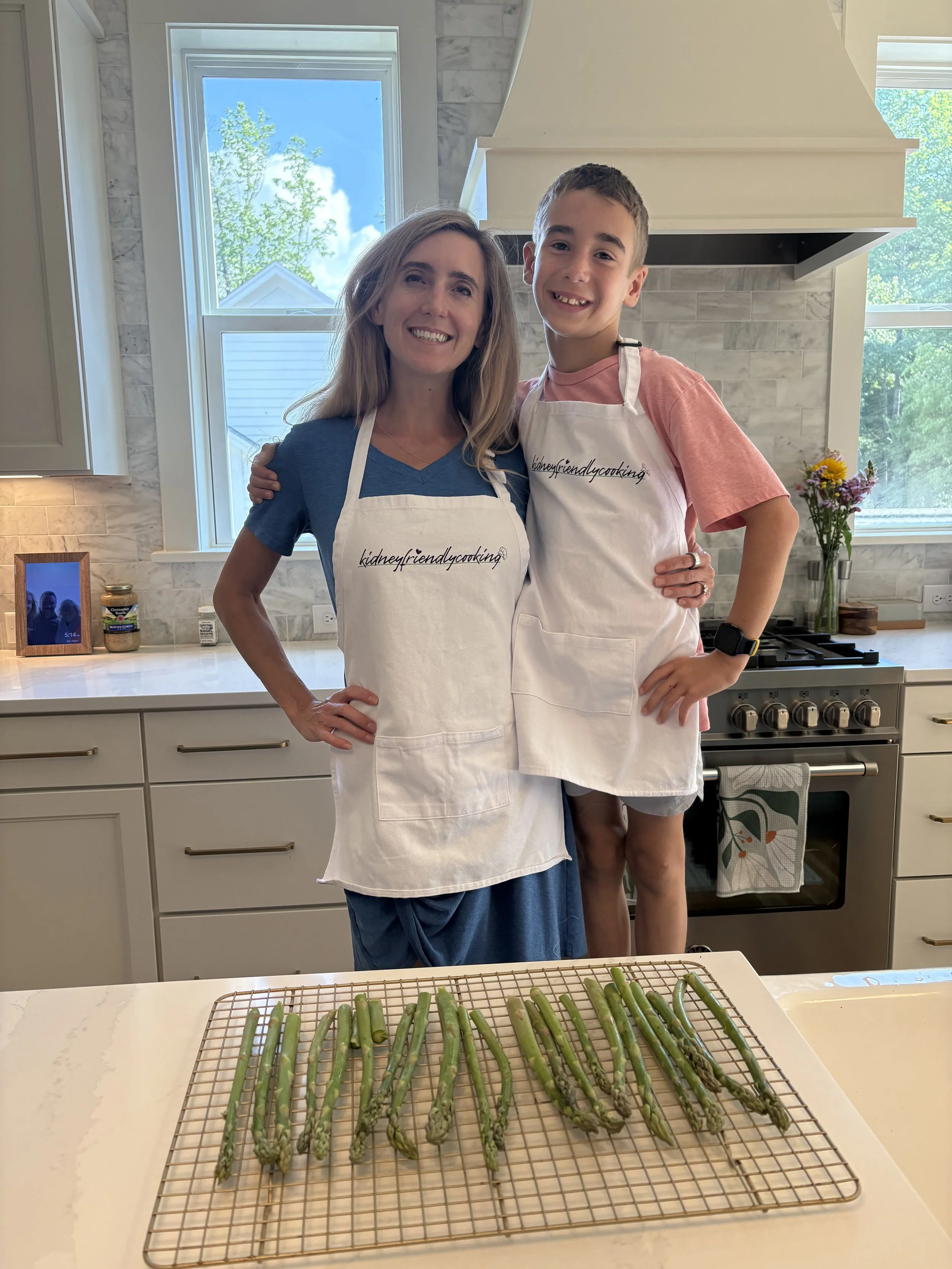 Two people, a woman and a boy, standing in a kitchen with a white countertop, smiling at the camera. They are wearing white aprons and appear to be preparing or drying asparagus, which is laid out on a wire rack on the countertop. The kitchen has white cabinets, a stove, and a vase with flowers on the counter. A large window shows a sunny day outside with blue sky and trees.