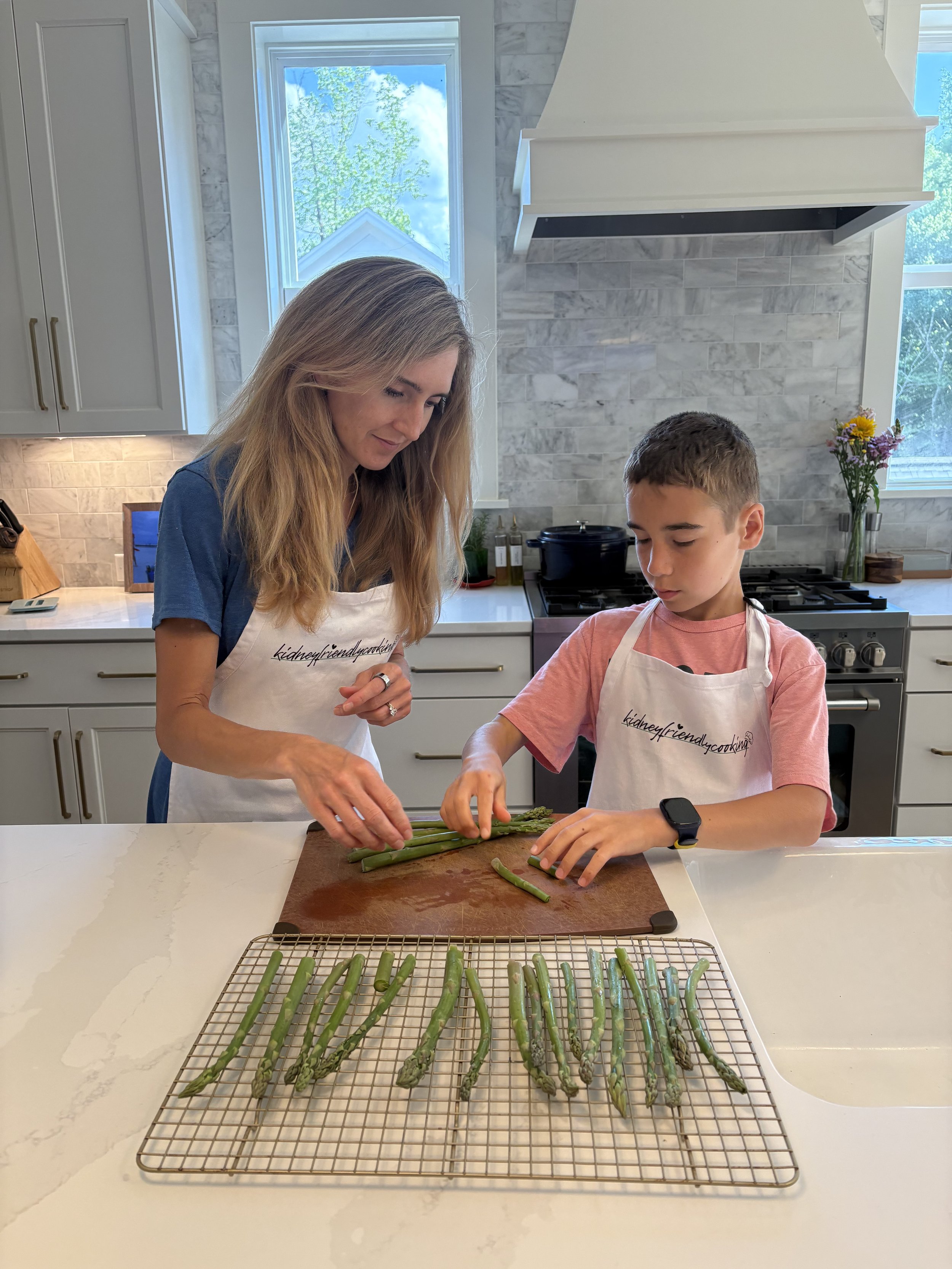 A woman and a young boy preparing asparagus in a modern kitchen with white cabinets, marble backsplash, and large windows. The asparagus is on a cooling rack on a white marble countertop.