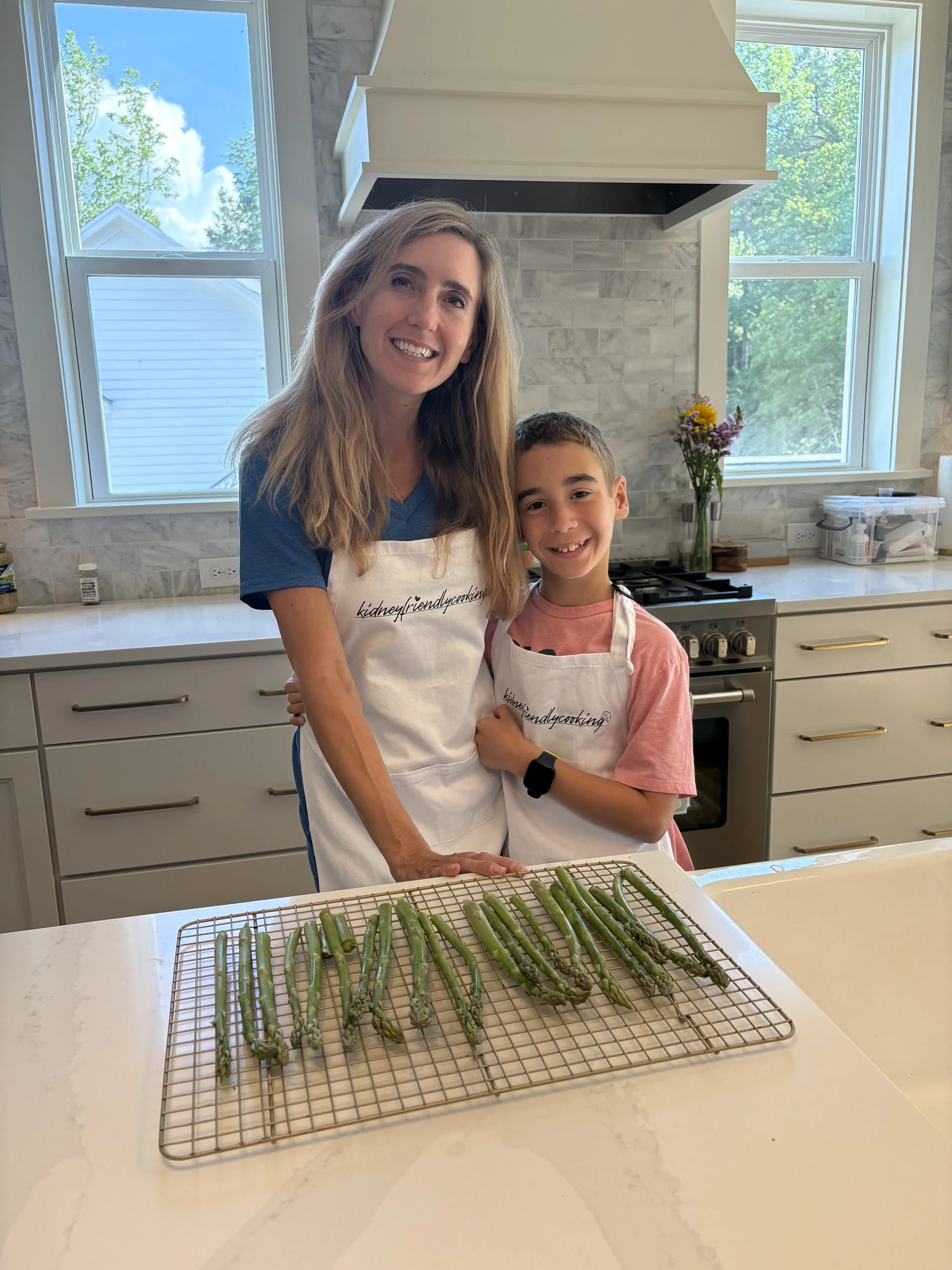 A woman and a young boy standing in a modern kitchen, wearing aprons, with a cooling rack of fresh asparagus on the white marble countertop in front of them. Large windows in the background show trees and a blue sky.