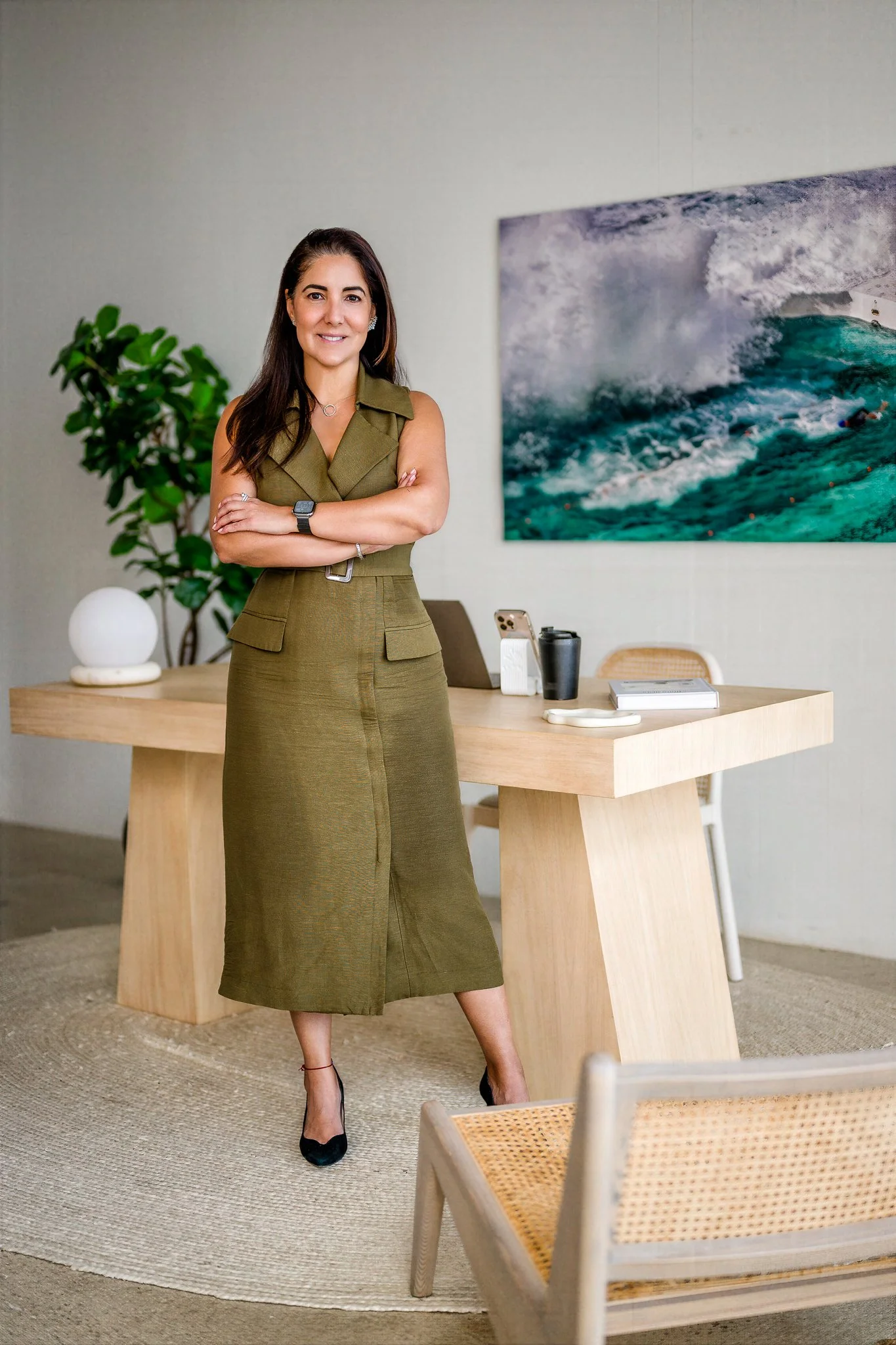 A woman in an olive green dress standing with arms crossed in an office space. Behind her is a large wall art of ocean waves, a wooden desk with a laptop, phone, and notebook, a white spherical lamp, and a green plant.