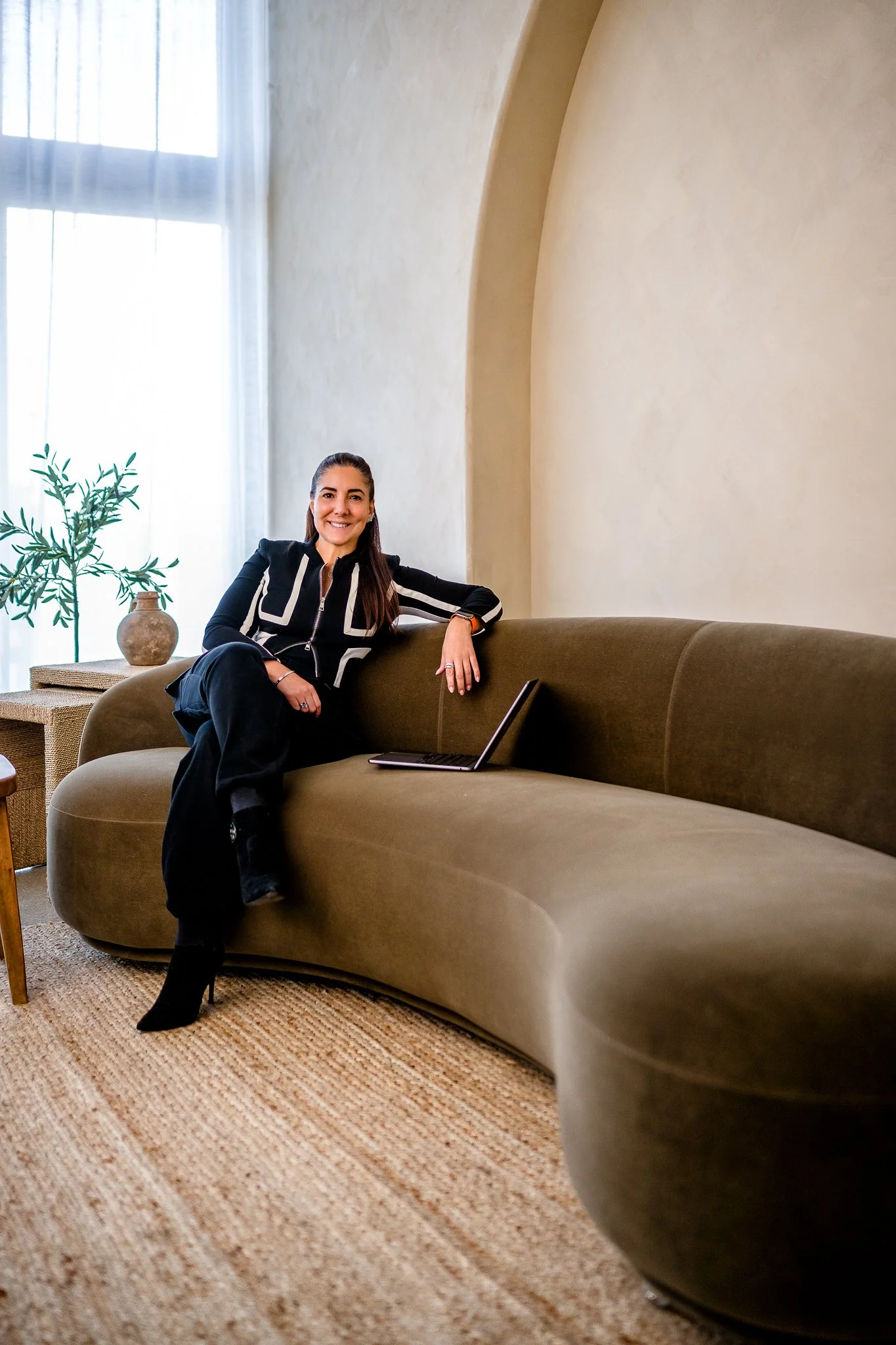 Smiling woman with long brown hair sitting on a green curved sofa in a well-lit room with a textured beige wall, a large window with sheer white curtains, a potted plant, and a laptop placed on the sofa.