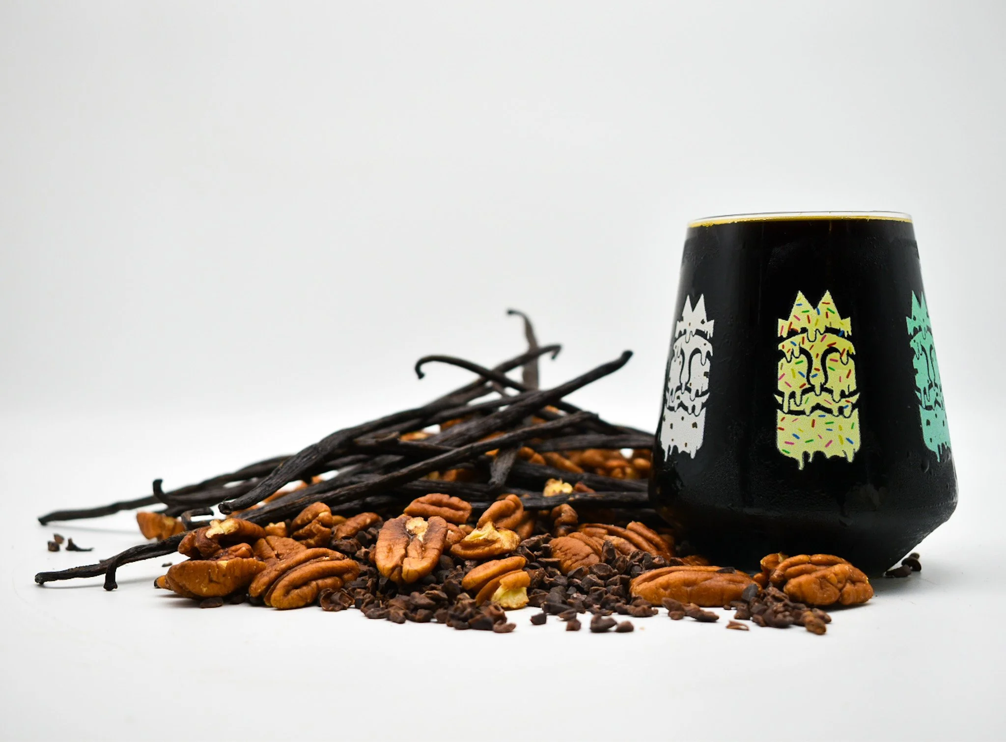 A black cup with colorful tiki masks design, surrounded by vanilla beans, chopped pecans, and chocolate chips on a white background.