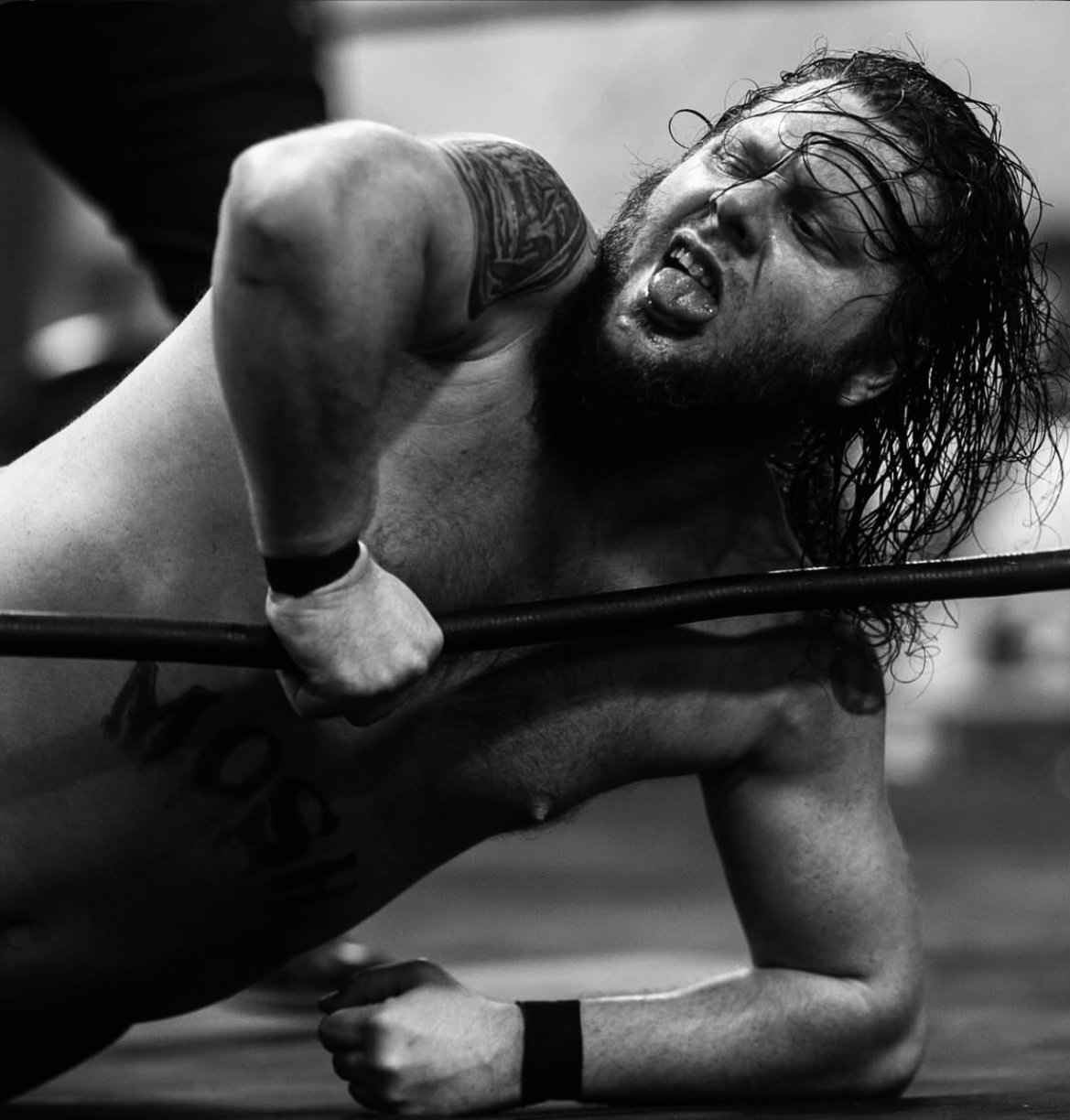 Black and white photo of a male wrestler on the mat, showing intense effort, with long wet hair, a beard, and a tattoo on his shoulder, holding a wrestling mat or bar.