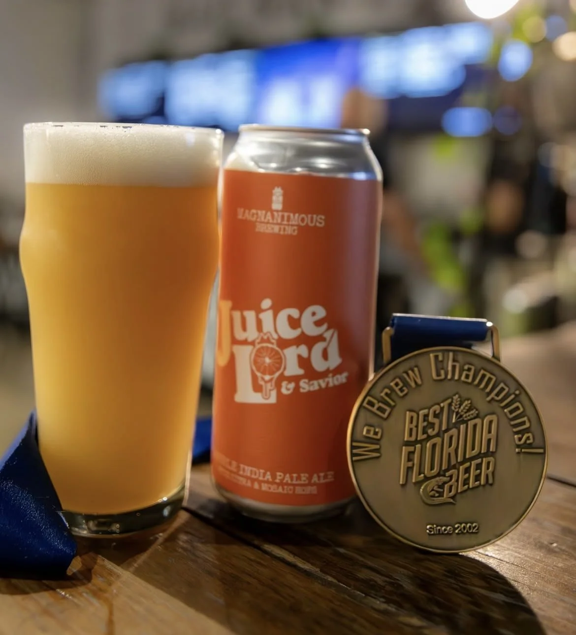 A glass of hazy IPA with a foamy head, a can of Juice Lord & Savior beer, and a gold medal with the inscription 'We Brew Champions! Best Florida Beer, Since 2002' on a wooden surface.