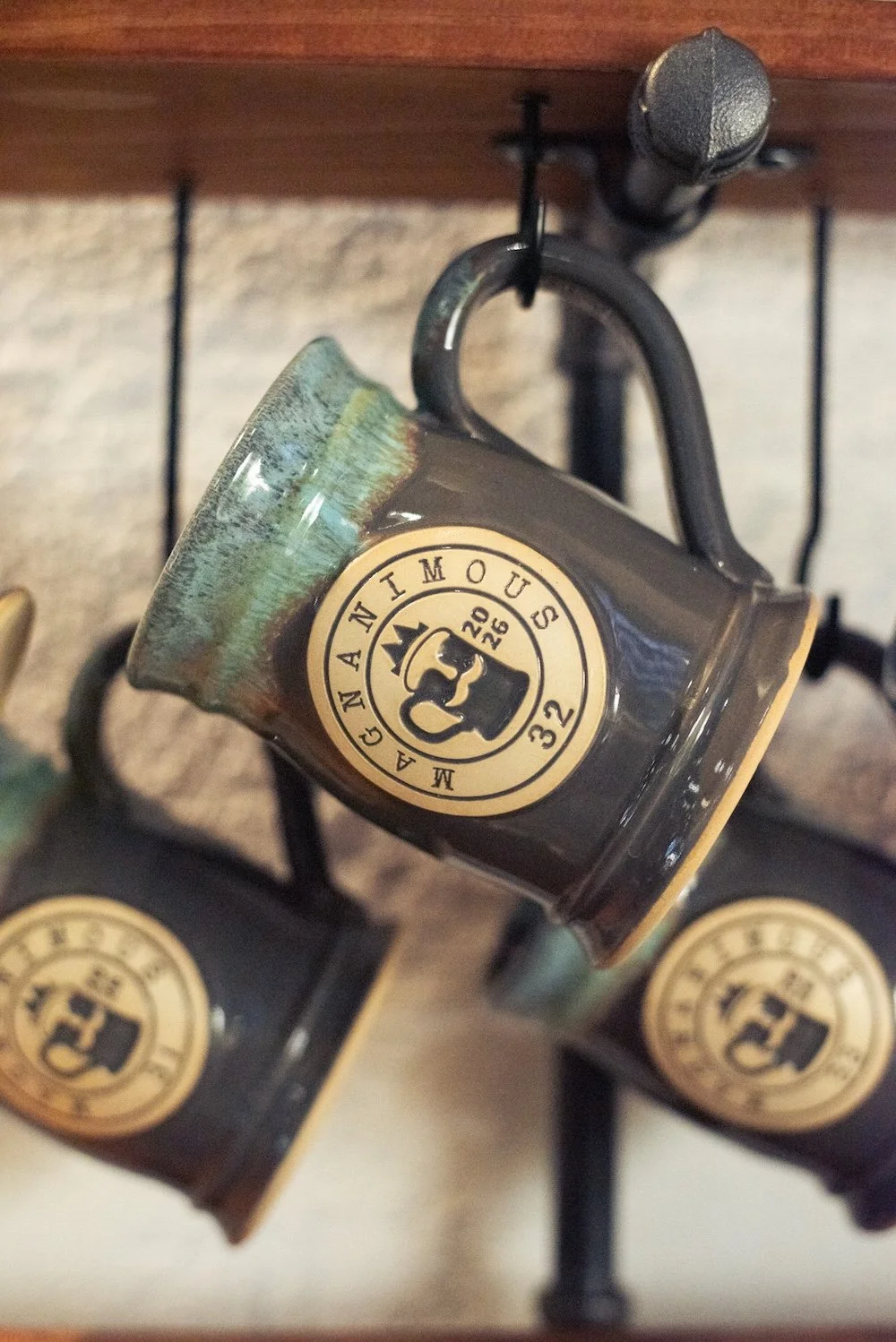 Close-up of black coffee mugs hanging from hooks on a wooden shelf, each mug features a logo with a crown and the text "MANCANIMOU" around the circle.