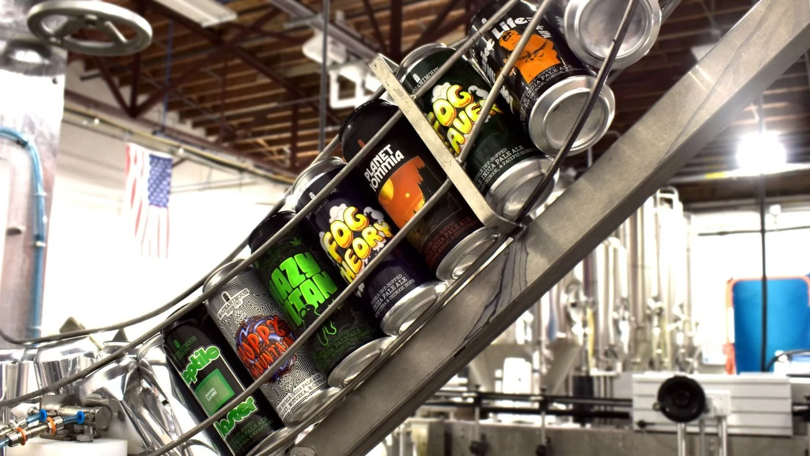 Galvanized metal conveyor belt carrying cans of craft soda with various labels, inside an industrial facility with exposed wooden ceiling beams and equipment in the background.