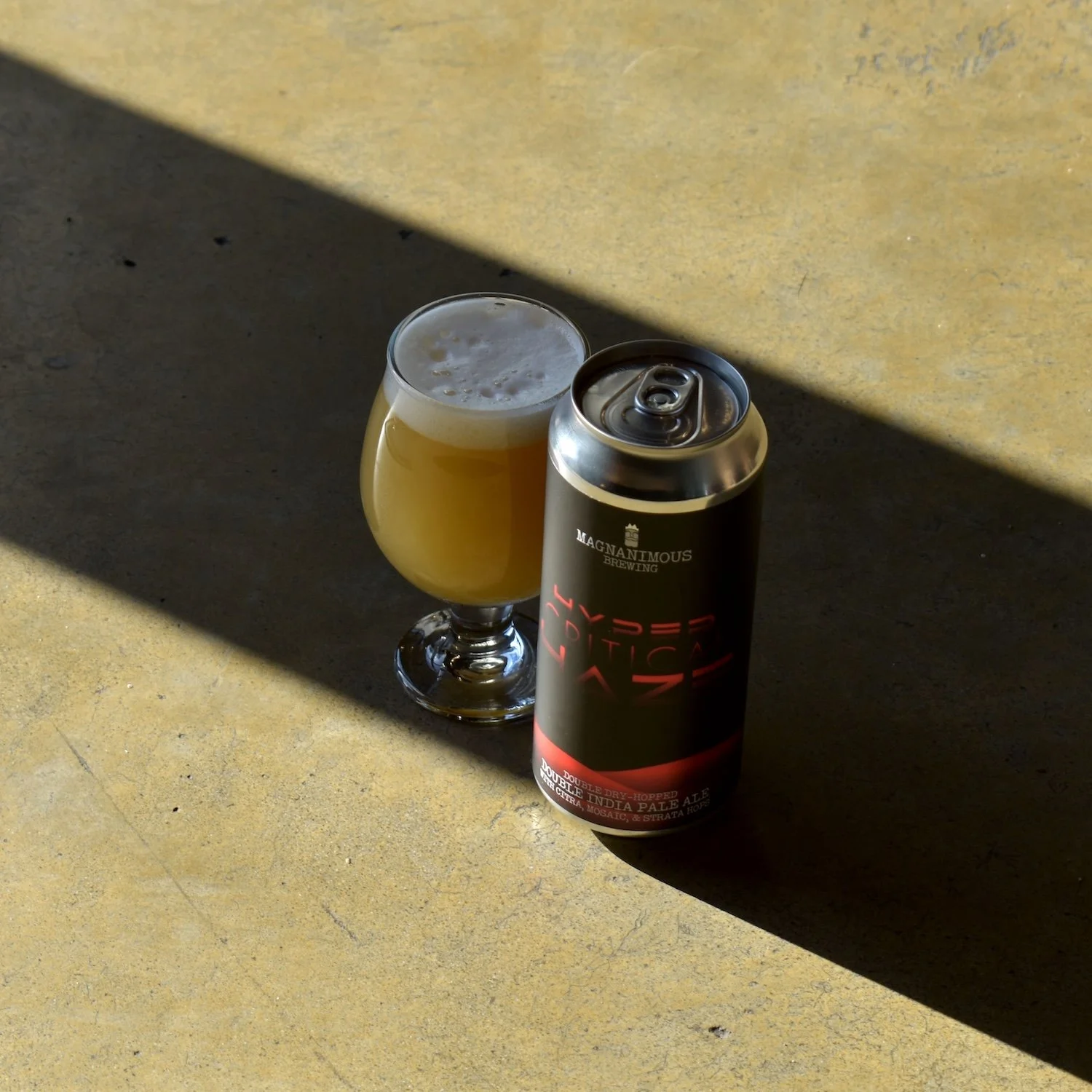 A glass of pale beer with a foamy head next to a black can of beer on a yellowish surface, with strong sunlight casting shadows.