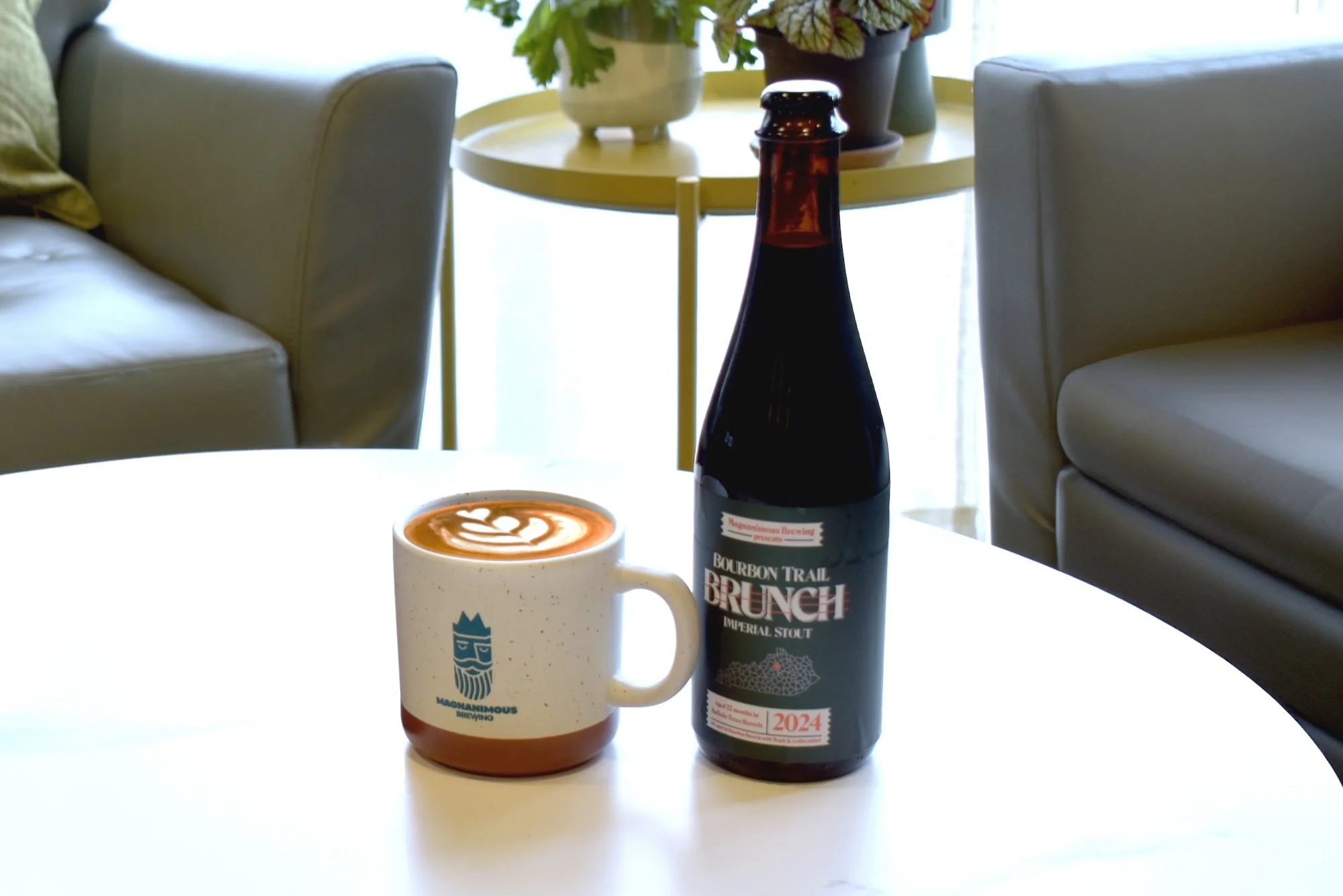 A white coffee mug with a logo of a stylized face and the words 'Magnanimous Brewing' on it, filled with a latte art coffee, placed on a white table. Next to it is a dark bottle labeled 'Bourbon Trail Brunch Imperial Stout 2024'. In the background, there are beige chairs and a yellow side table with plants.