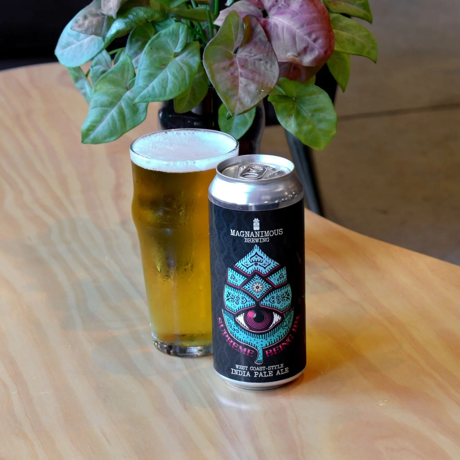 A tall glass of golden beer with a frothy head and a can of Magnanimous Brewing's Extreme Behemoth India Pale Ale next to it, placed on a wooden table with a potted plant with green and purple leaves in the background.