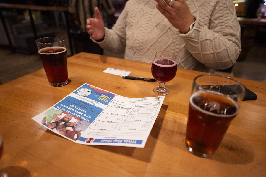 Two glasses of dark soda on a wooden table, with a flyer, a pen, a glass of red wine, and a person's hands in a beige sweater visible in the background.