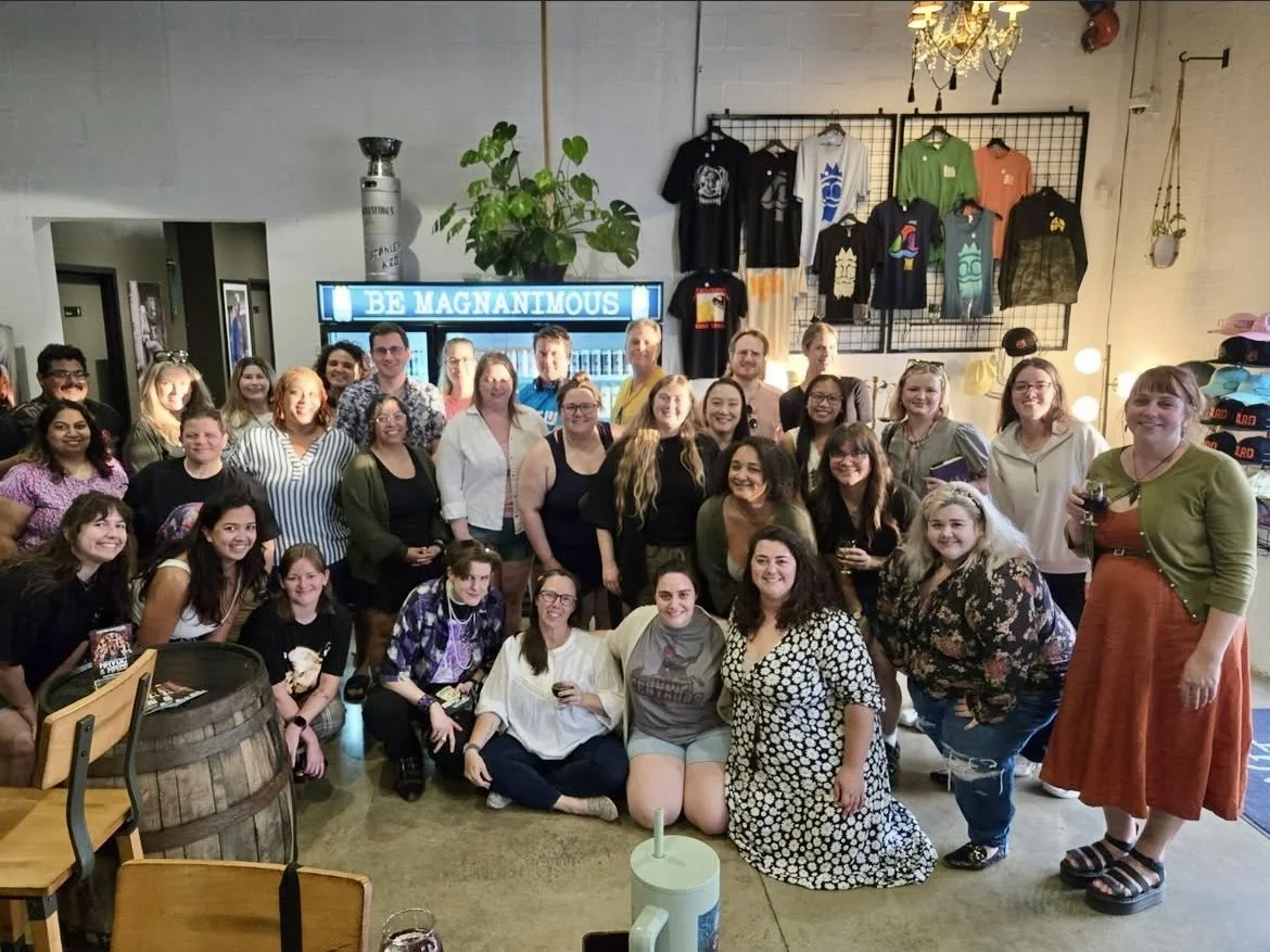 Group of people gathered inside a retail store or cafe, posing for a photo with clothing and merchandise displayed on the walls behind them. Some are holding drinks, and there is a sign that reads 'BE MAGNANIMOUS' in the background.