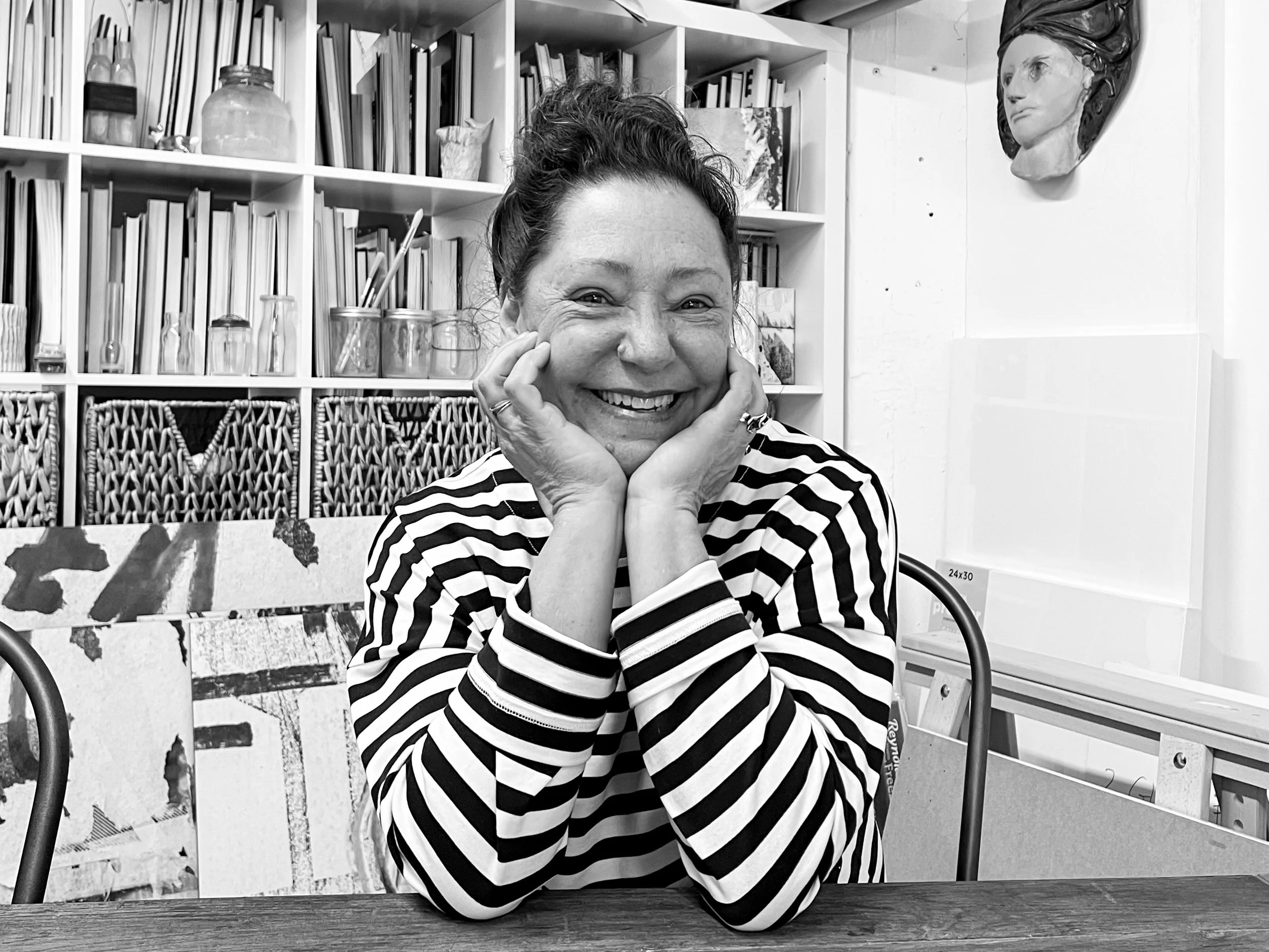 A woman with a big smile, resting her face on her hands, wearing a striped shirt, indoors with a background of bookshelves and art on the wall.