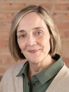 A woman with short blonde hair and a gentle smile, wearing a green shirt and beige sweater, standing against a brick wall background.