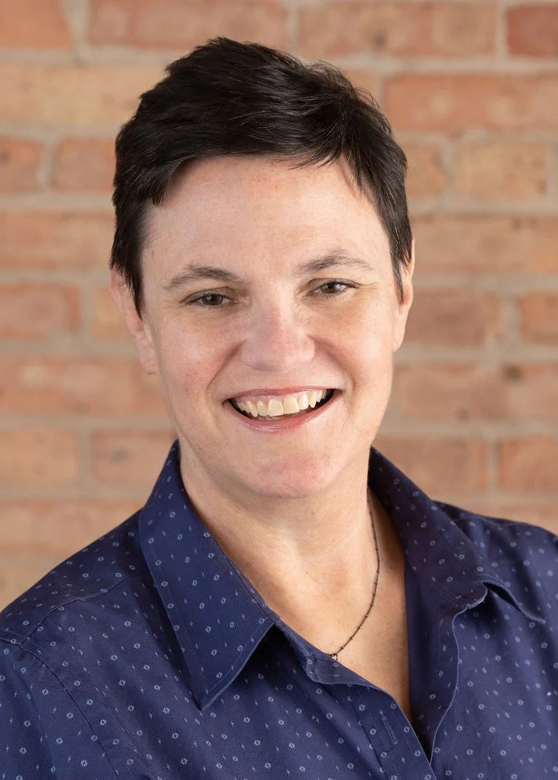 A person with short dark hair, smiling, wearing a navy blue shirt with a subtle pattern, and a thin necklace, standing in front of a brick wall.