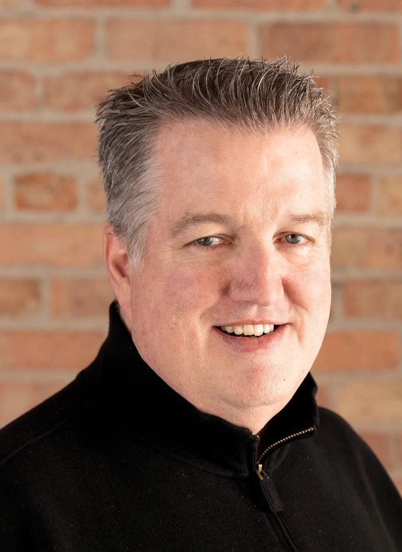 Portrait of a smiling middle-aged man with short, grayish hair, wearing a black zip-up sweater, standing in front of a brick wall.
