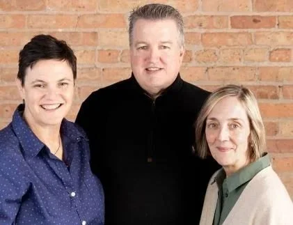 Three people standing in front of a brick wall, smiling at the camera.