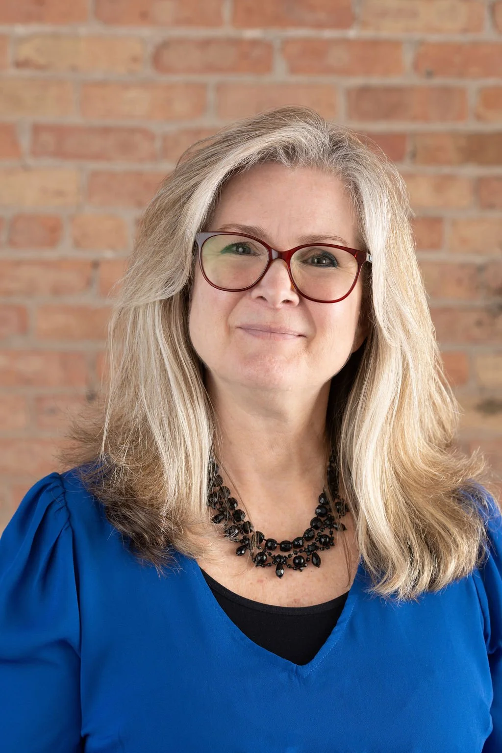 A woman with blonde hair, wearing red glasses, a blue top, and a black necklace, smiling in front of a brick wall.