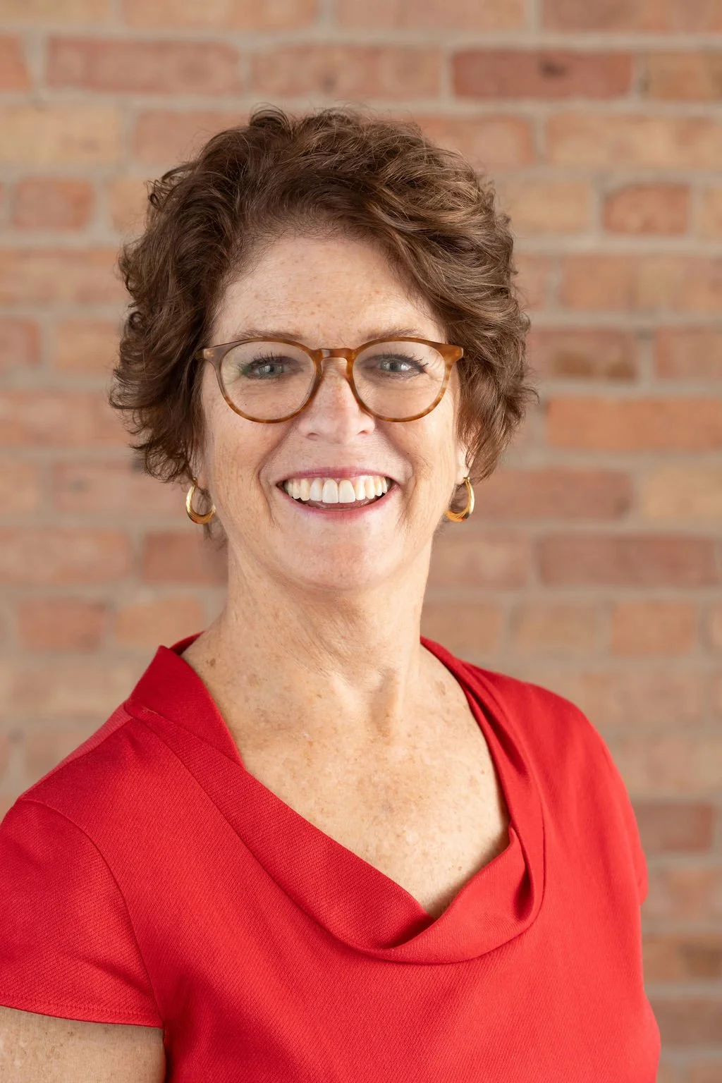 A woman with short curly brown hair, wearing glasses and gold hoop earrings, smiling at the camera against a brick wall background. She is wearing a red top.