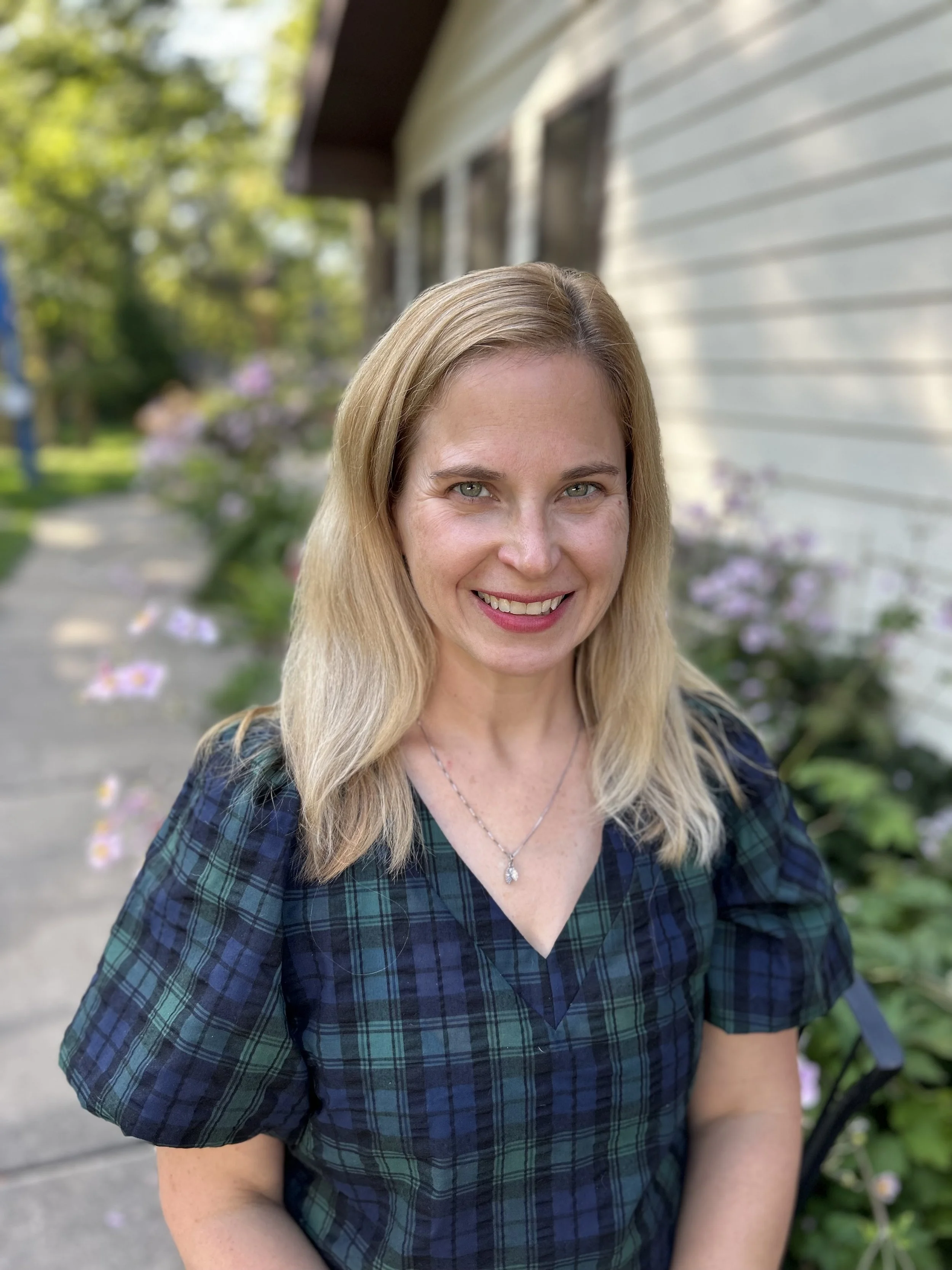 A woman with blonde hair smiling outdoors in front of a house with white siding and purple flowers.