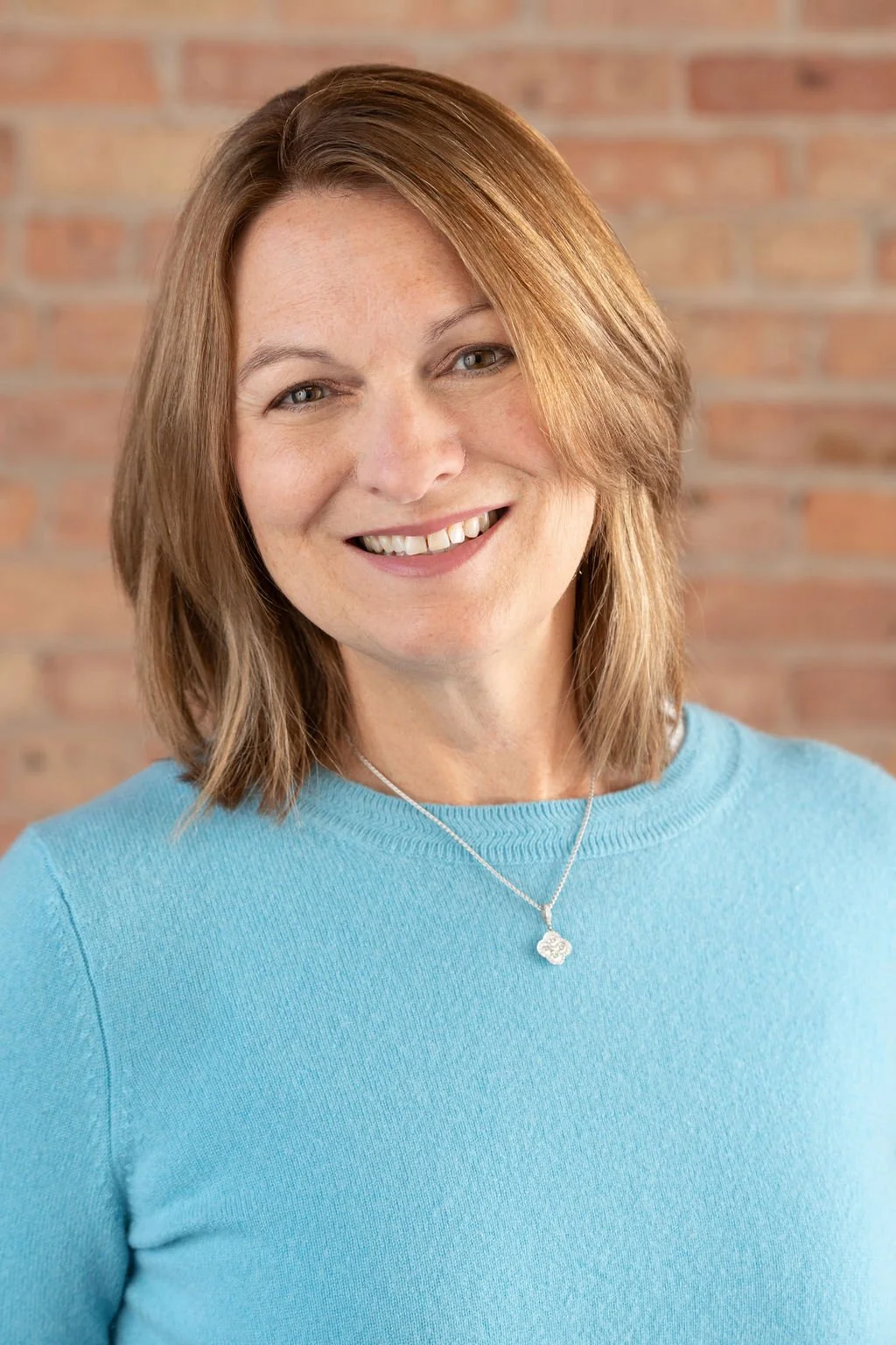 A woman with shoulder-length light brown hair, wearing a light blue sweater and a necklace, smiling in front of a brick wall background.