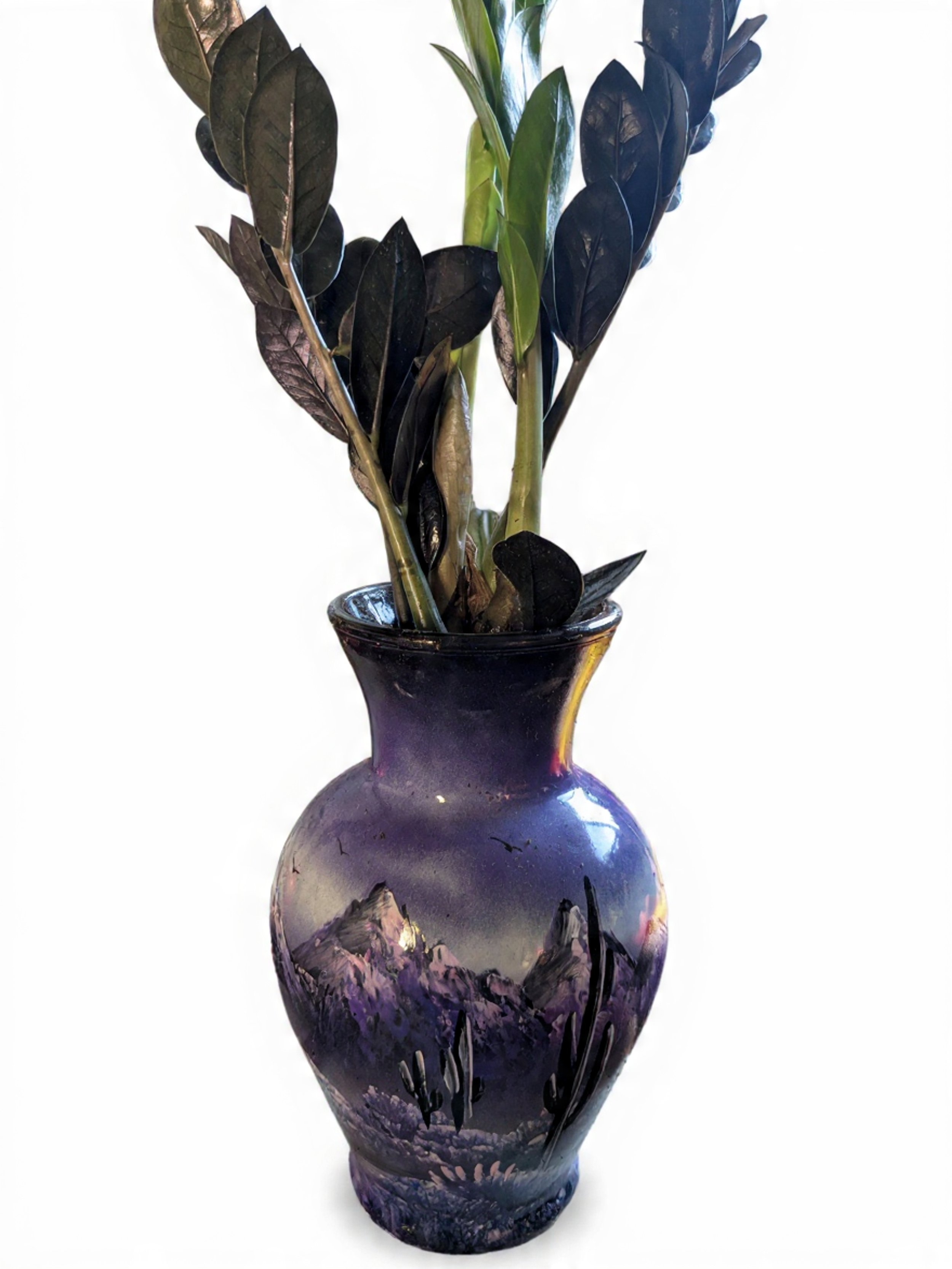 A decorative vase with a desert mountain landscape painted on it, holding a branch of dark green and brown leaves.