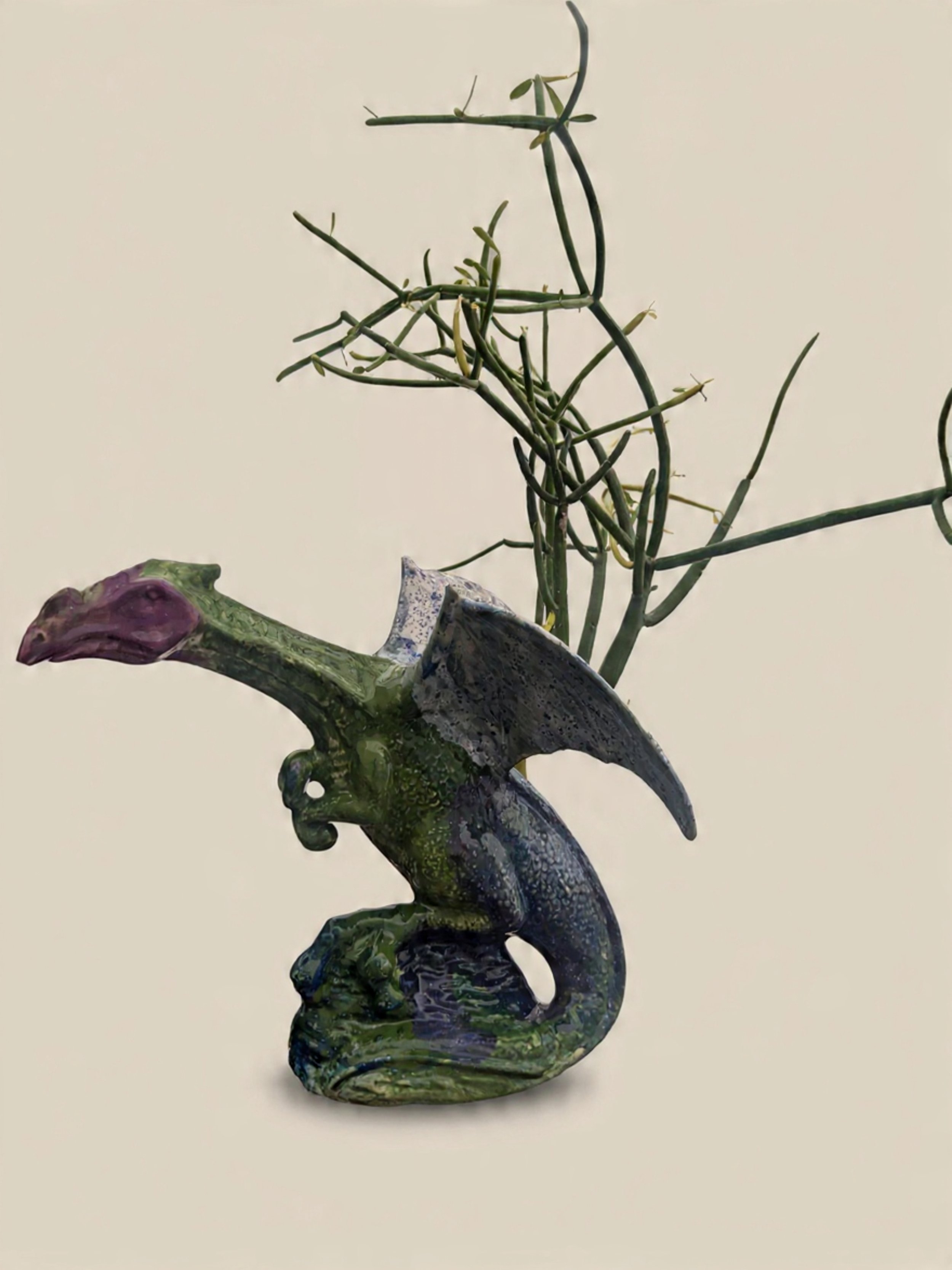 A decorative dragon-shaped sculpture with wings, green and purple coloring, and a crab clasped in its tail, with a tall plant with multiple thin, branching stems in the background.