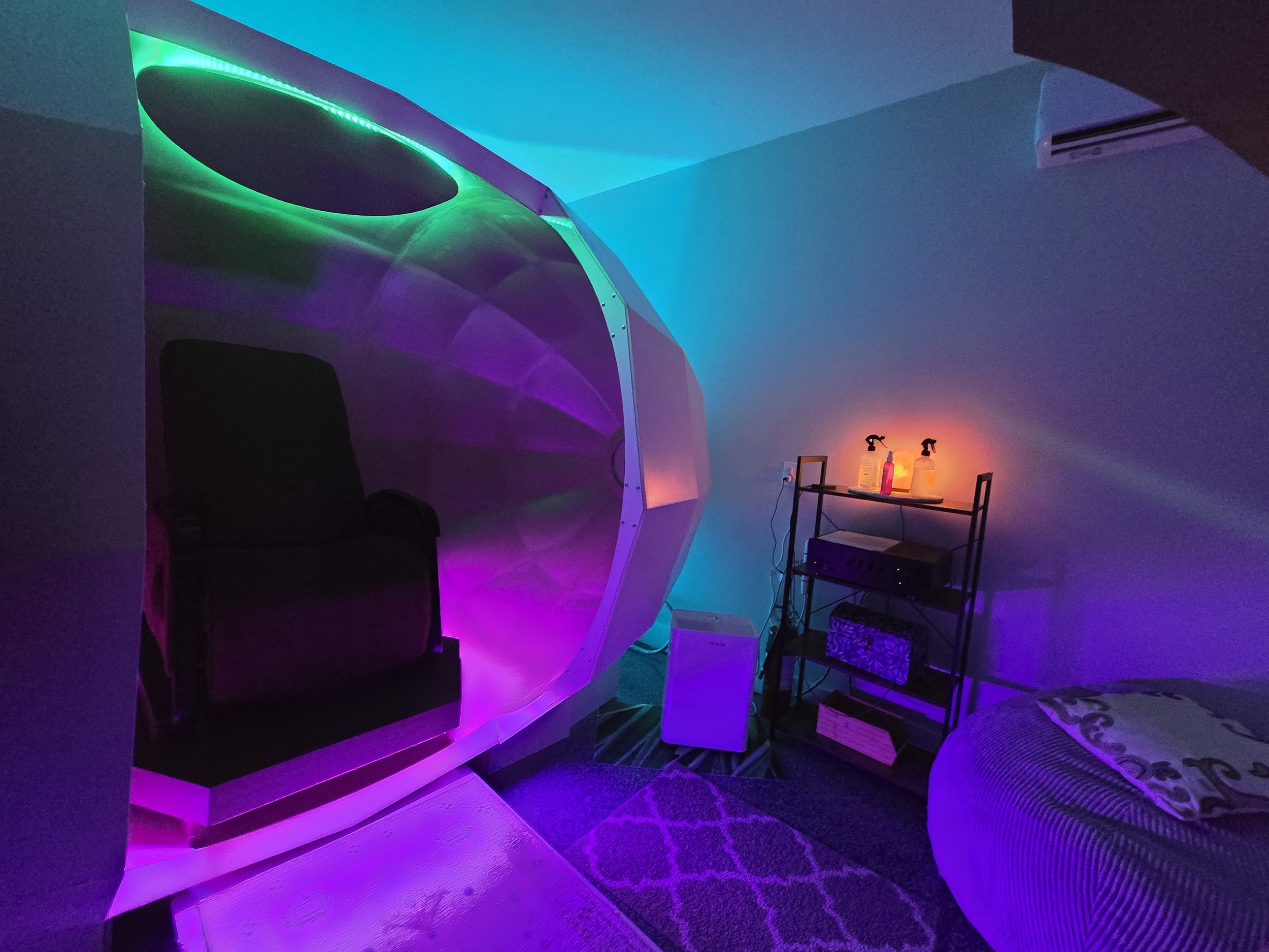 A room with glow lighting and a modern enclosed pod or capsule with colorful LED lights. There is a chair inside the pod, a bedside table with spray bottles, a small shelf with electronic equipment, a framed picture, a blanket on the bed, and a pink patterned rug.