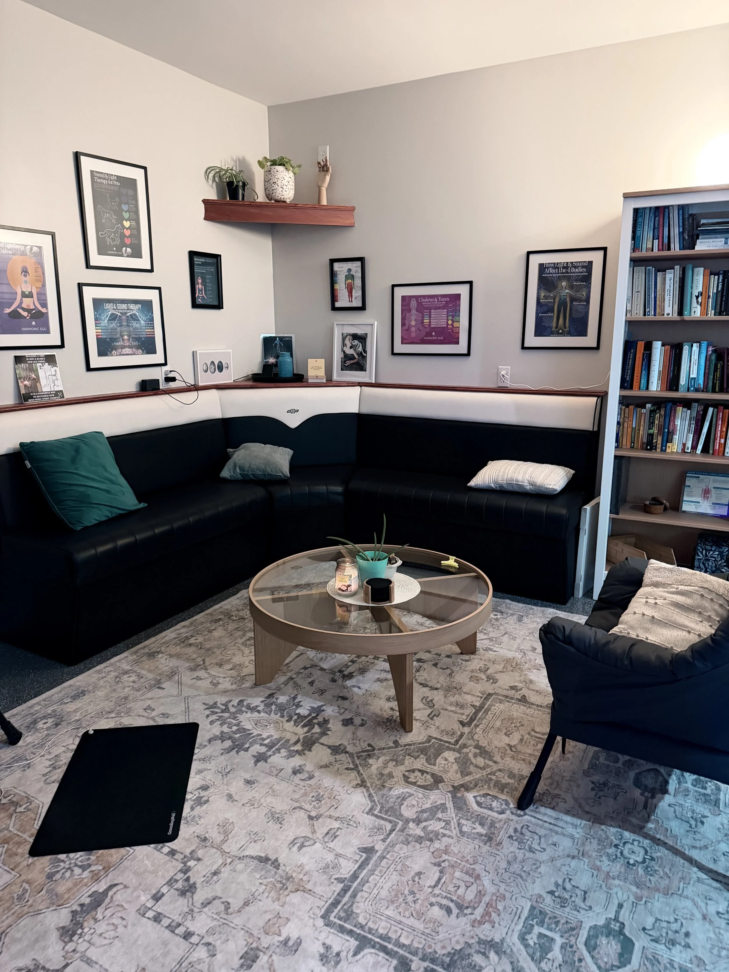 A cozy waiting room with black leather sofa, a wooden coffee table with plants and candles, framed artwork and posters on the wall, a bookshelf filled with books, and a black armchair with a blanket. There's a patterned rug on the floor and a tablet device near the sofa.