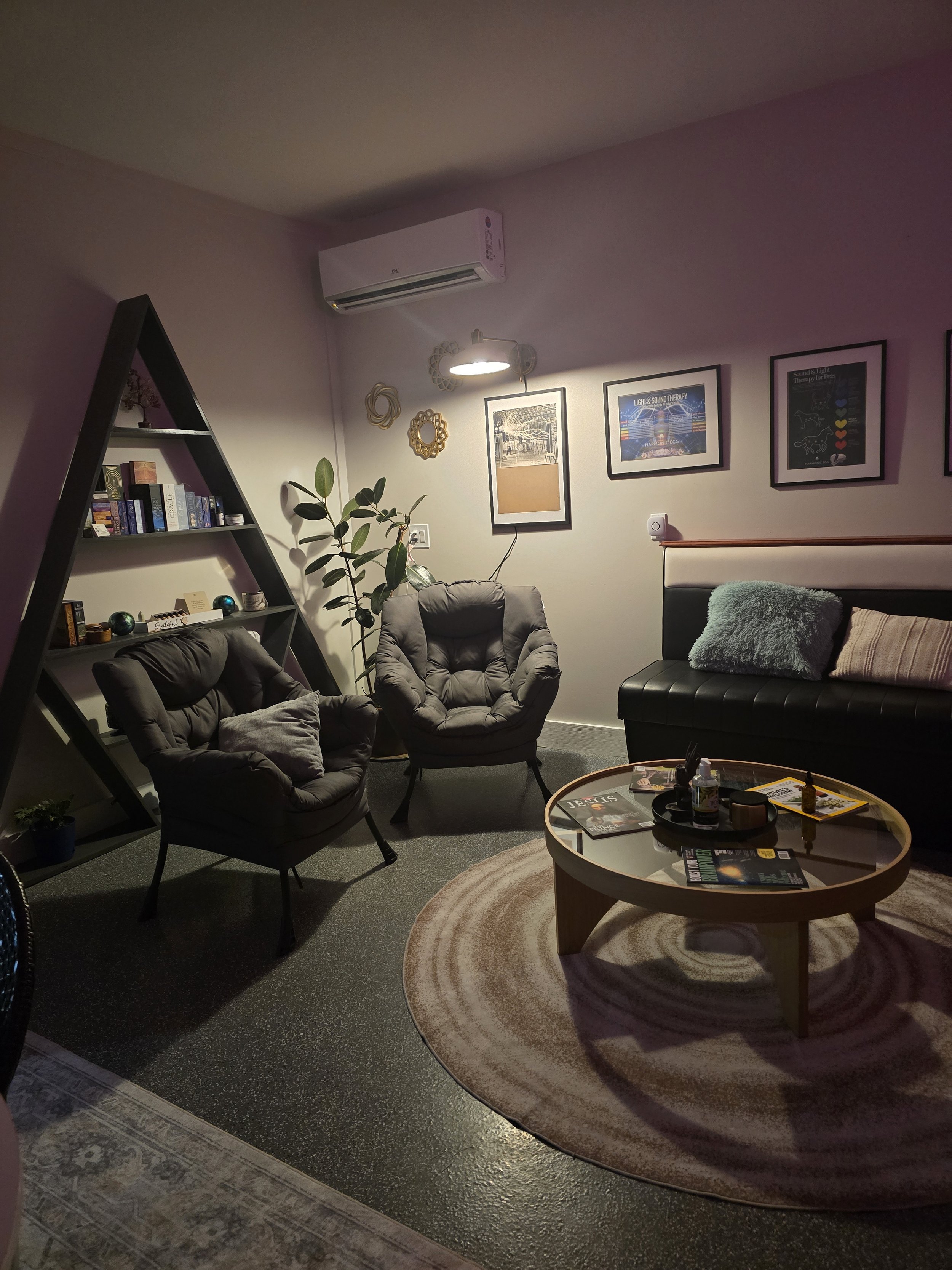 Cozy living room with two armchairs, a sectional sofa with pillows, a round coffee table with magazines, a bookshelf shaped like a triangle, framed pictures on the wall, and a potted plant, illuminated by a reading lamp and ceiling light.