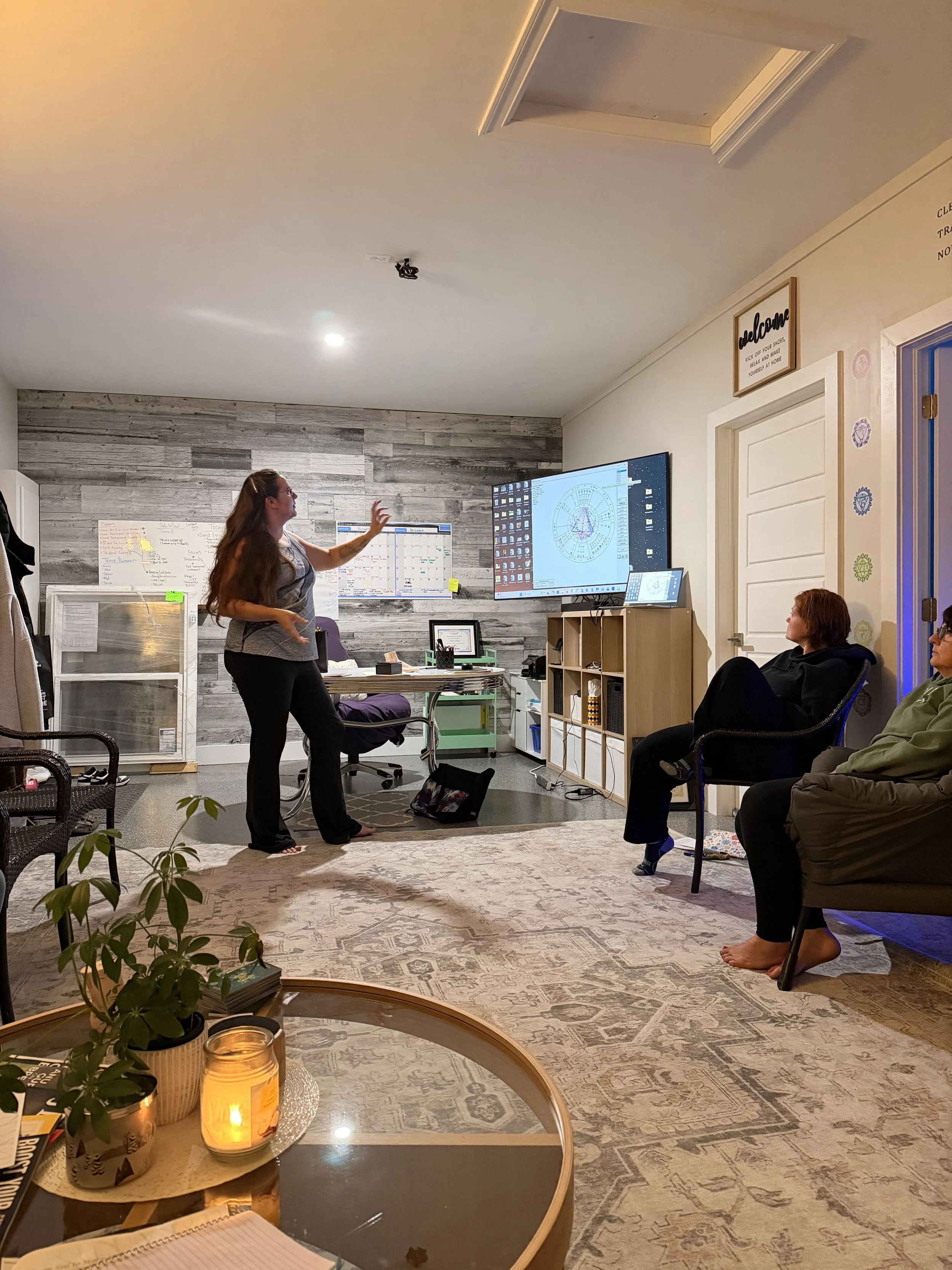 A woman stands and gestures while giving a presentation in a cozy room with three seated women listening. The room has a large wall-mounted screen, shelves, and a whiteboard. A rug covers the floor, and there is a table with a plant, candle, and notebooks in the foreground.