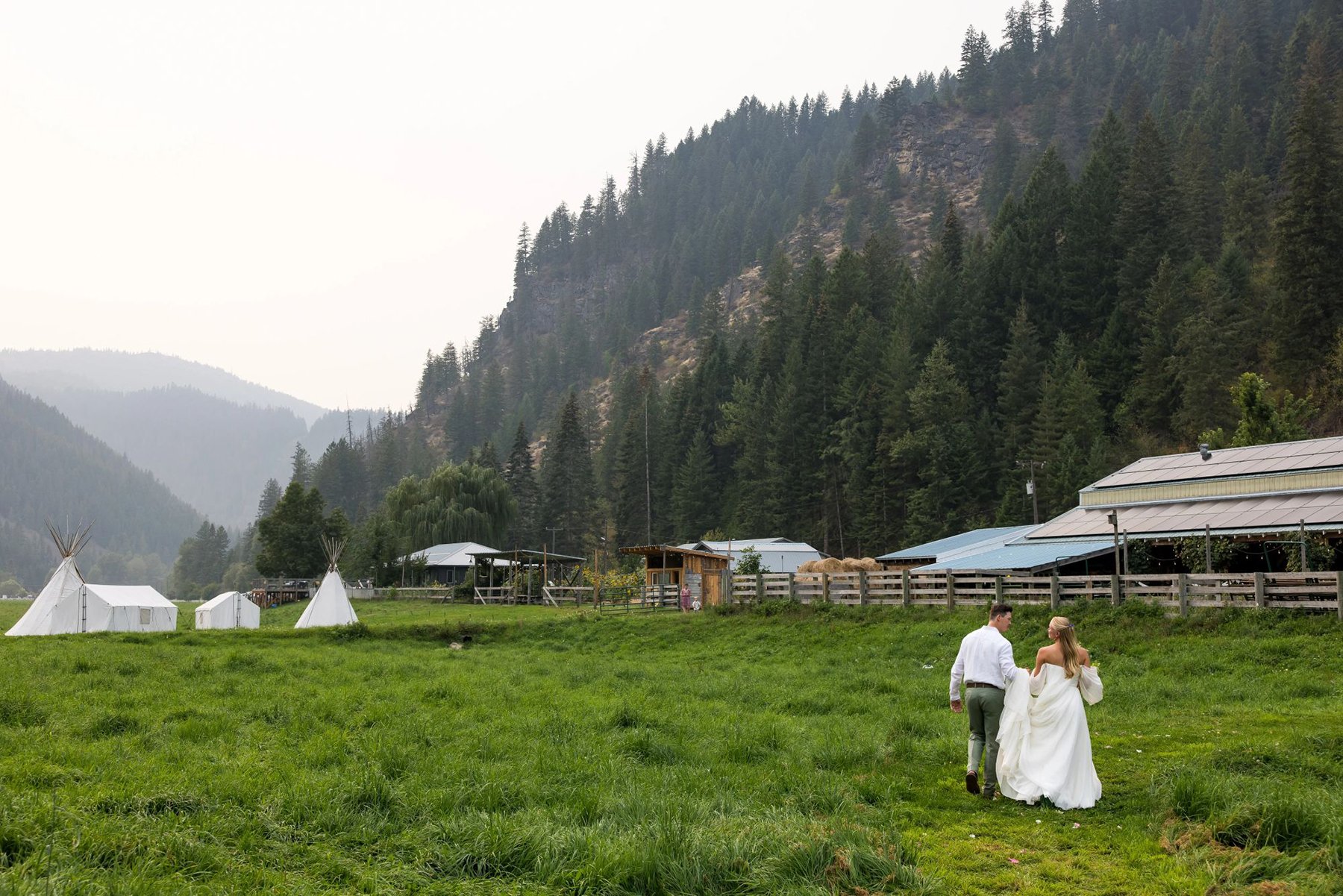 A bride and groom walking hand-in-hand across a grassy field with teepees and farm buildings in the background, surrounded by forested mountains.