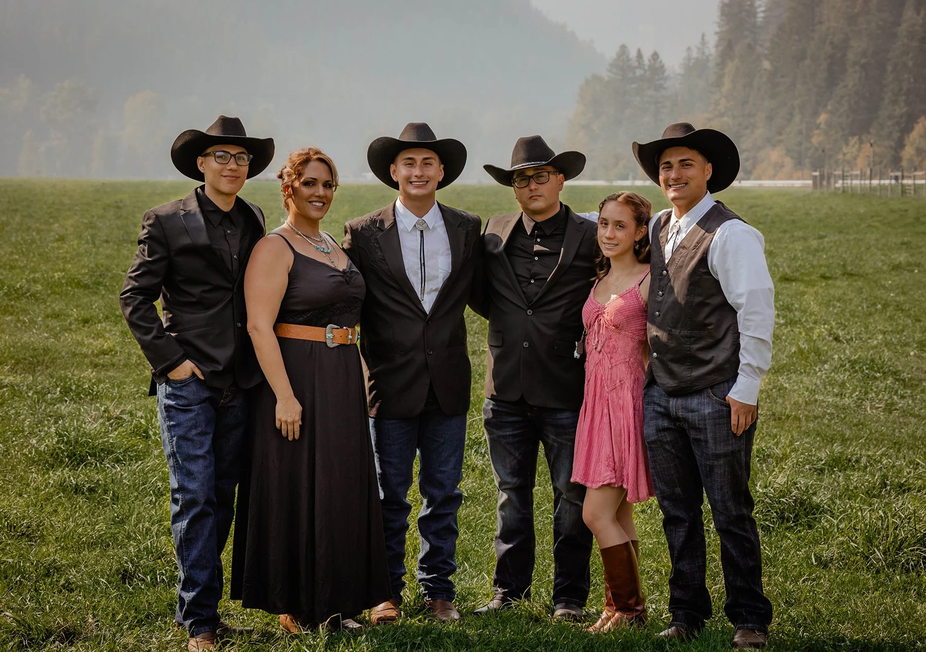 Group of six people in western outfits standing on a grassy field with forest and mountains in the background.