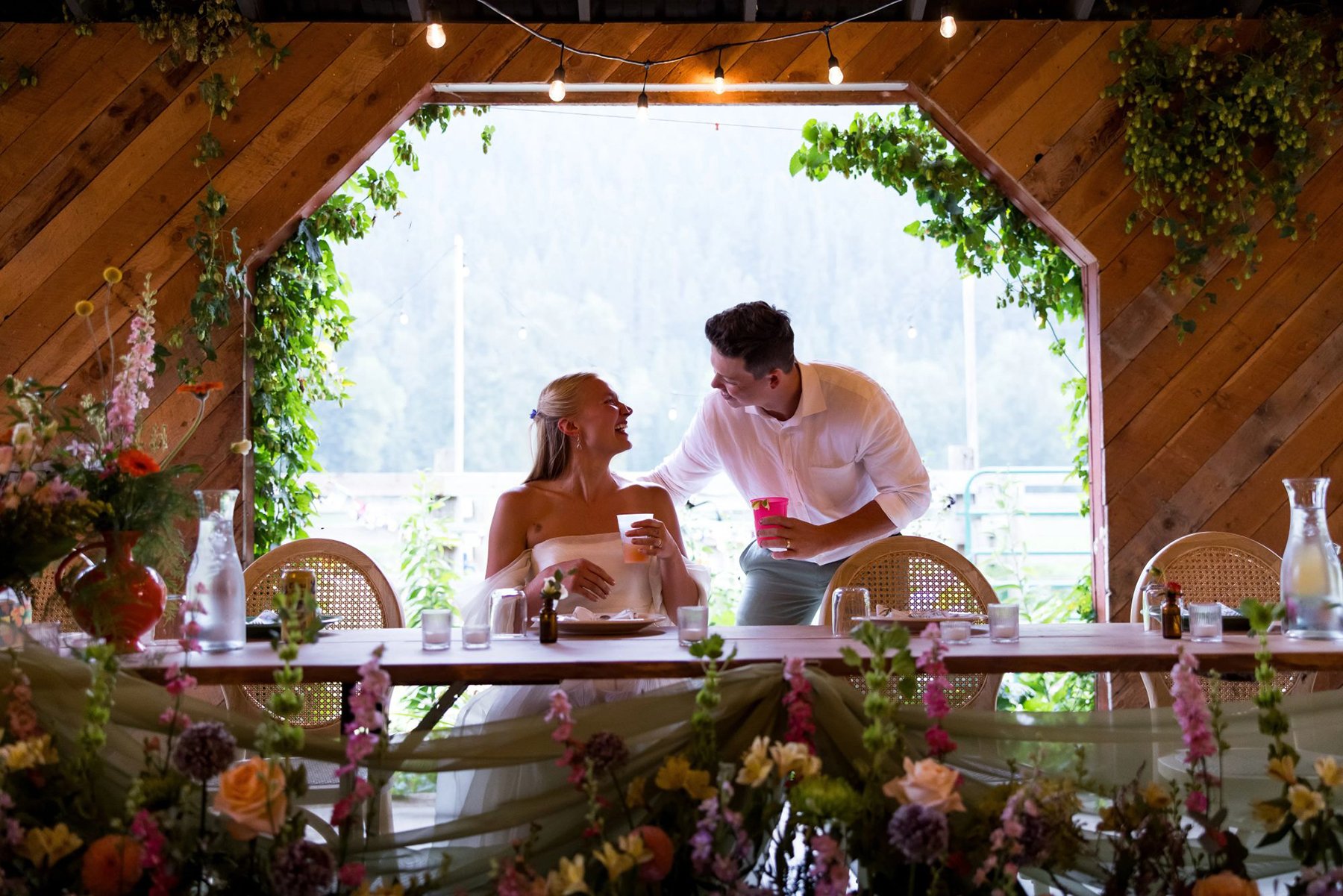 A bride and groom at a wedding reception, sitting at a table decorated with flowers, with the groom offering a drink to the bride inside a venue with wooden walls and string lights.