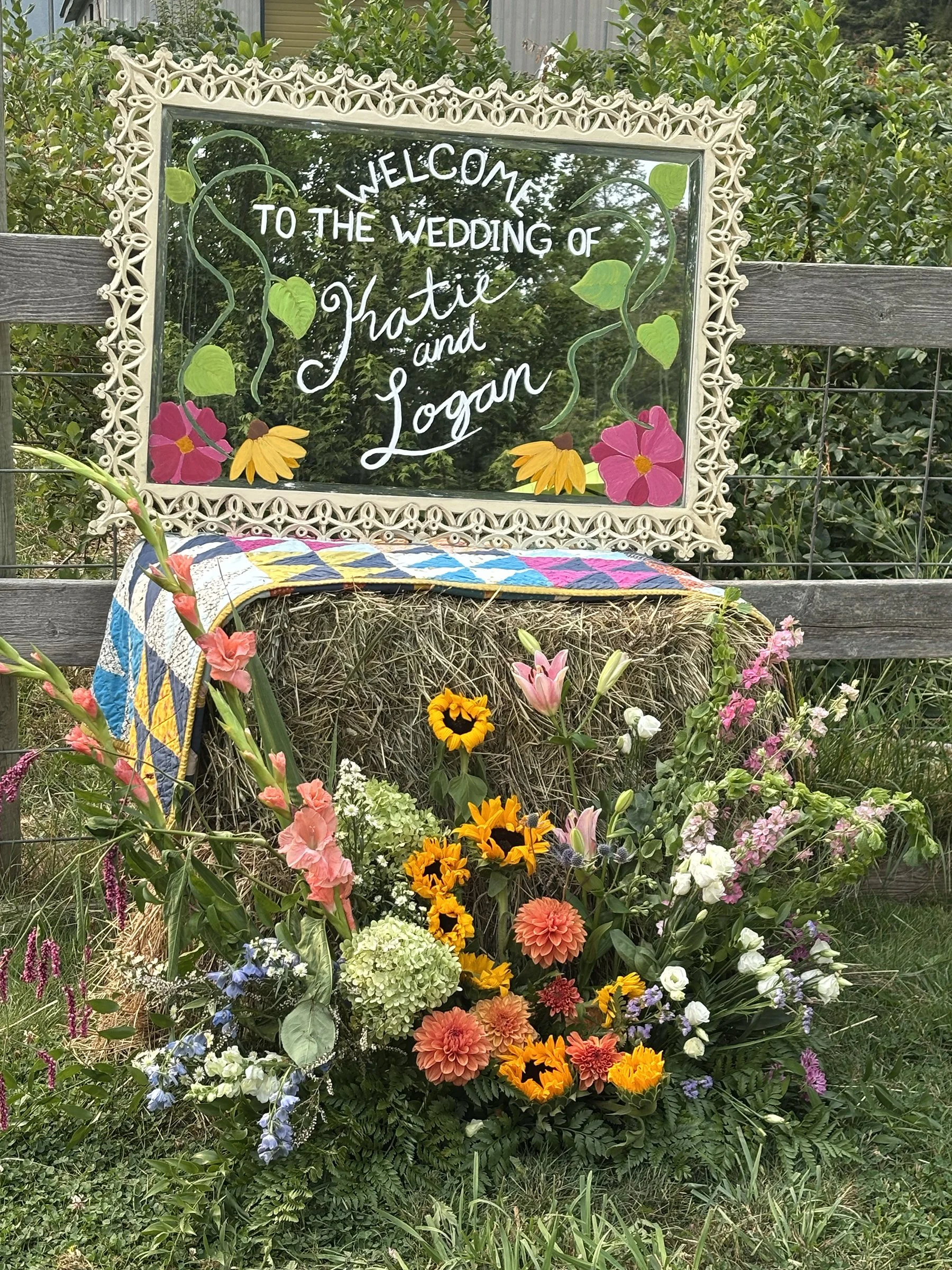 Signboard with a decorative white frame reading "Welcome to the wedding of Katie and Logan" in white lettering, surrounded by green leaves and pink flowers painted on the glass. The sign is placed behind a hay bale covered with a colorful quilt, with