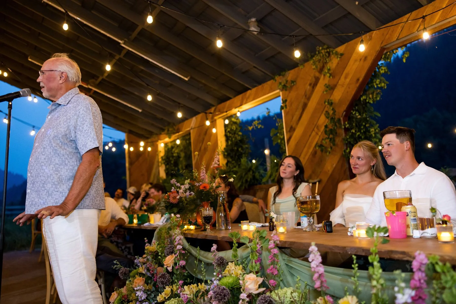 An elderly man giving a speech at a wedding reception, with a wedding couple and guests sitting at a decorated table with flowers, candles, and drinks, under string lights and a wooden ceiling with greenery.