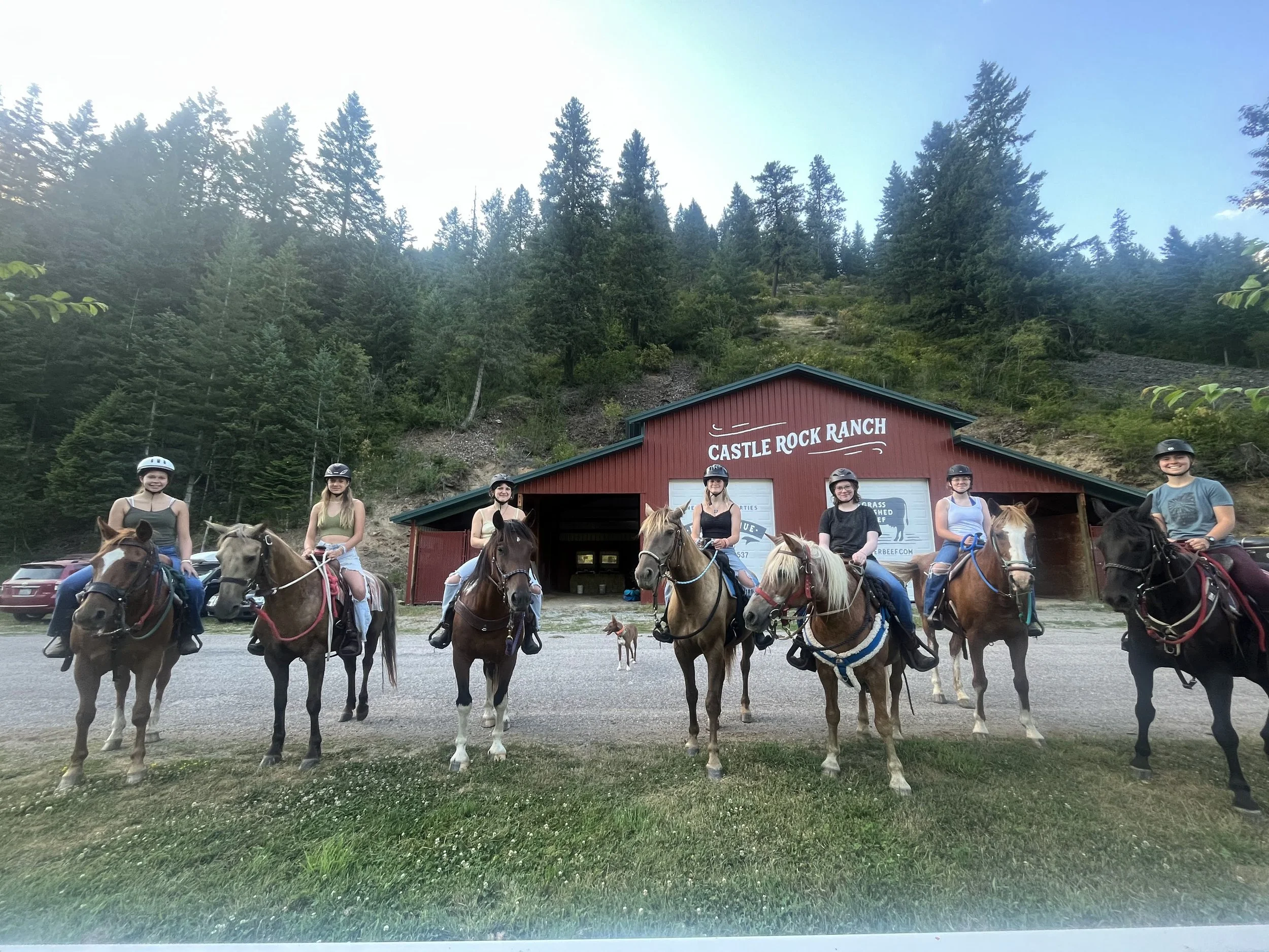 A group of seven people on horseback in front of a red barn labeled 'Castle Rock Ranch,' with a forested hillside in the background.