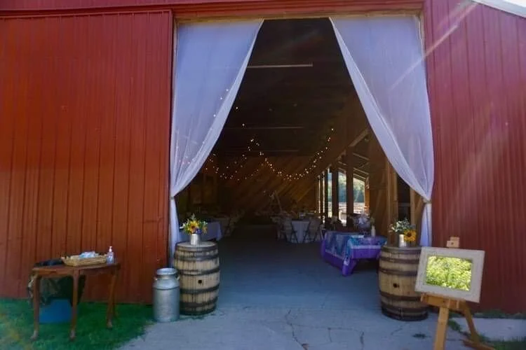 Entrance of a rustic barn converted into event space, with red wooden siding, white curtains, decorated tables, wine barrels, and string lights inside.