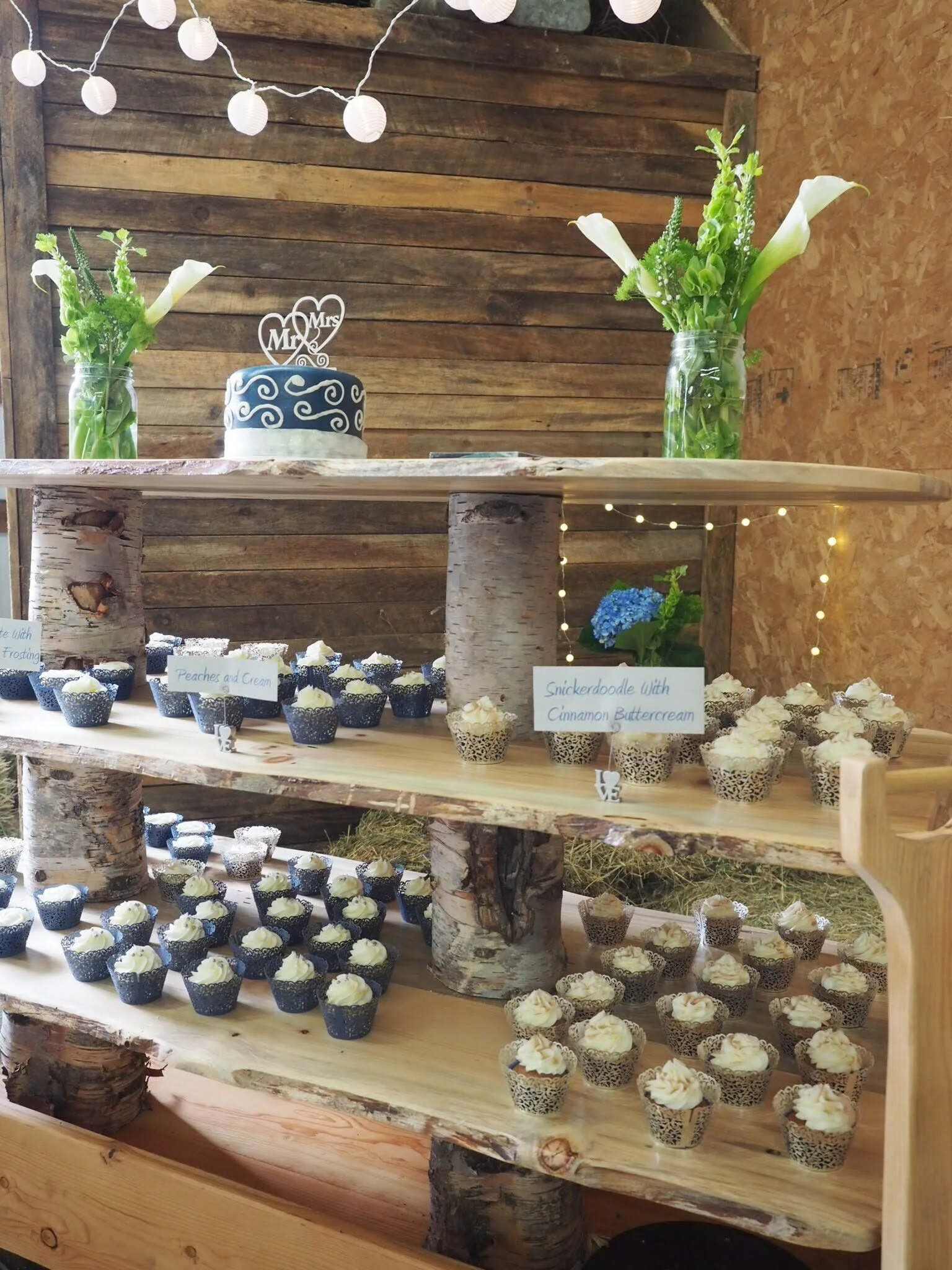 A rustic wooden dessert table decorated with a wedding cake, two vases of white calla lilies, and blue hydrangeas. Multiple cupcakes with white frosting and decorative wrappers are arranged on the table, with handwritten labels for different flavors.
