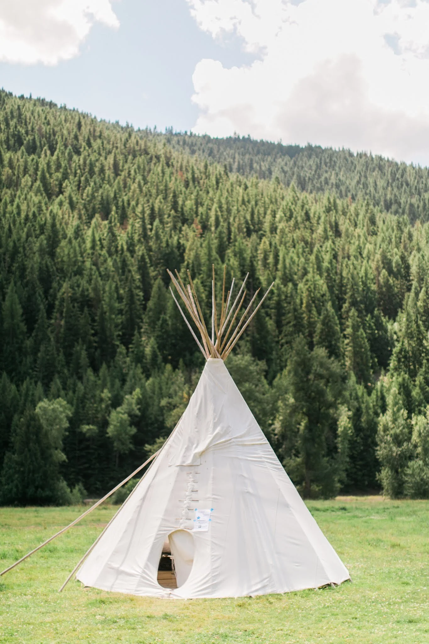 A white teepee tent on a grassy field with a forested mountain in the background and partly cloudy sky.