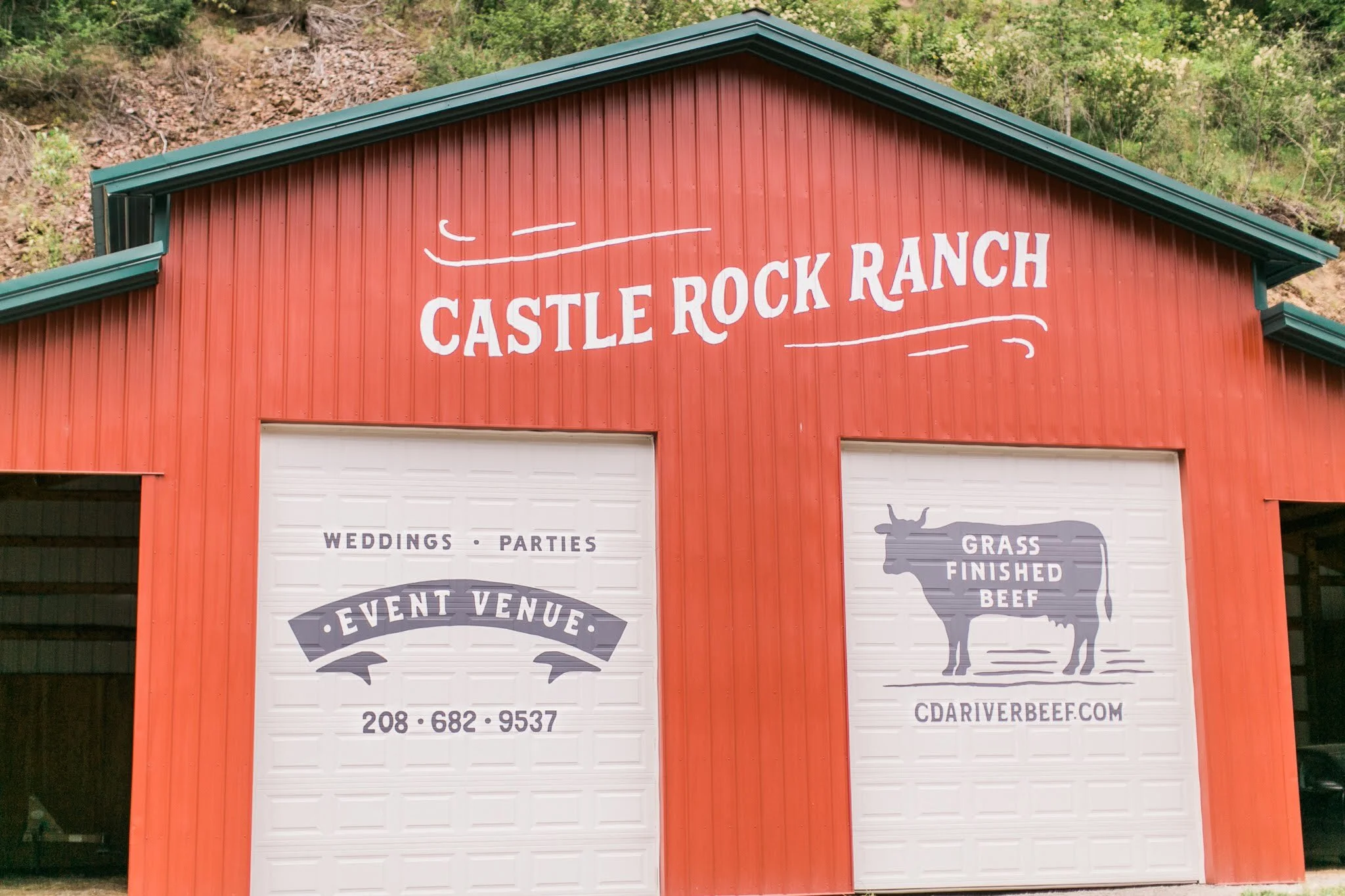 Red barn-style building with white lettering that reads 'Castle Rock Ranch'. Two white garage doors with black and gray graphics and text; one says 'Event Venue' and the other shows a cow silhouette with 'Grass Finished Beef' and the website 'CDriver