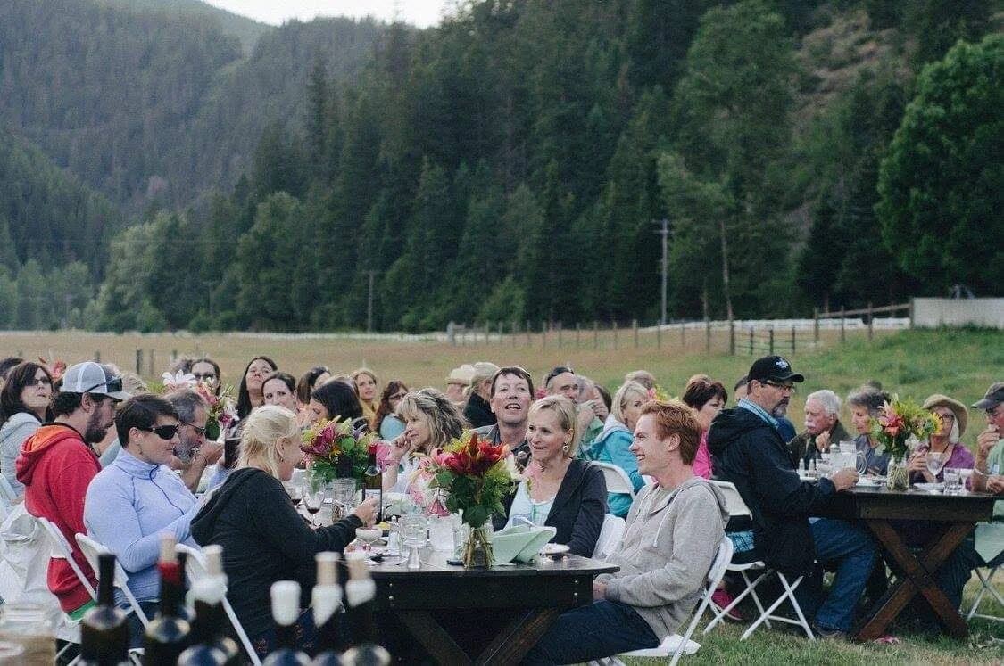 People sitting at long outdoor table with flowers, enjoying a gathering or celebration in a scenic rural area with trees and hills in the background.