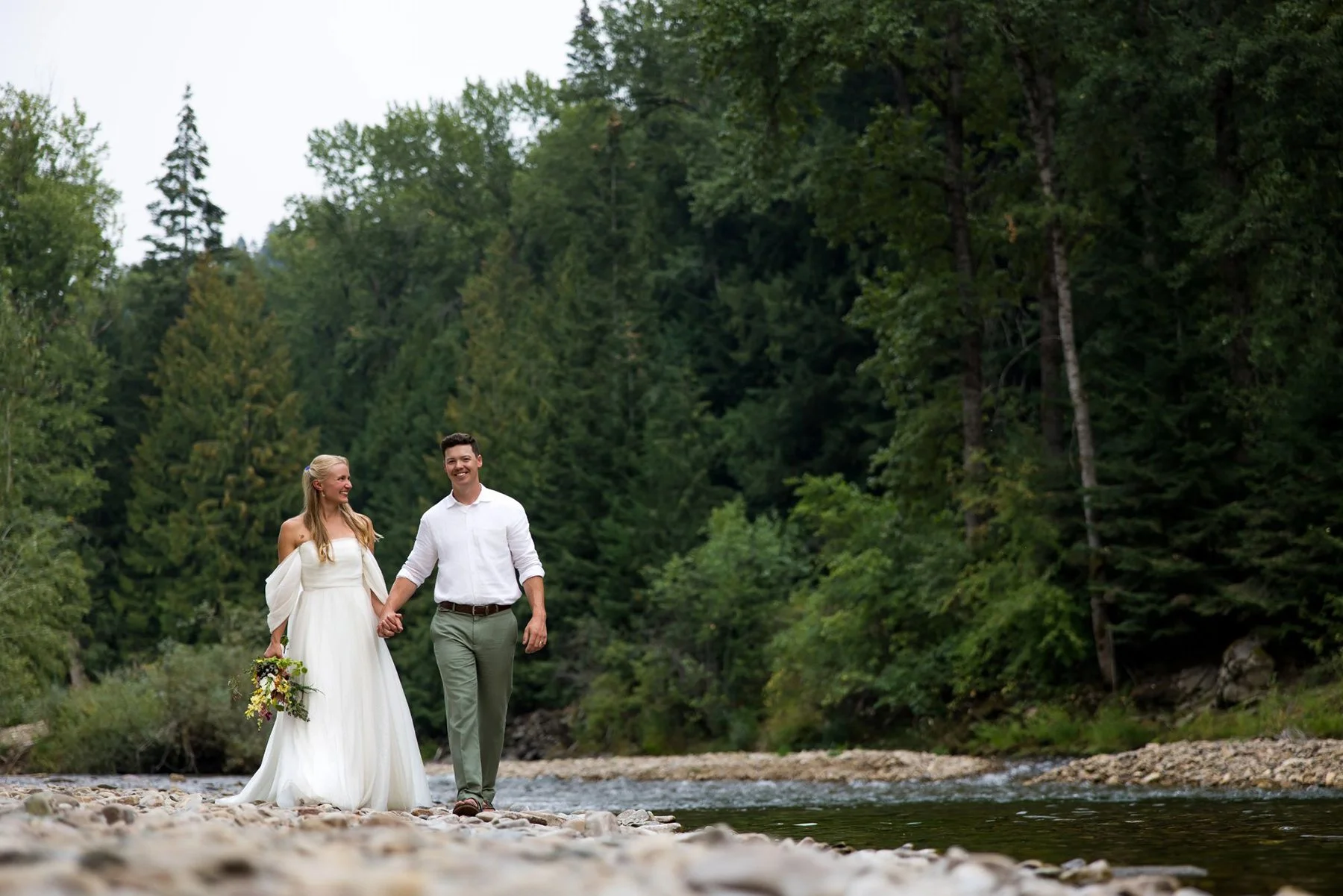 A newlywed couple walking hand in hand along a rocky riverbank surrounded by green trees, with mountains in the background.