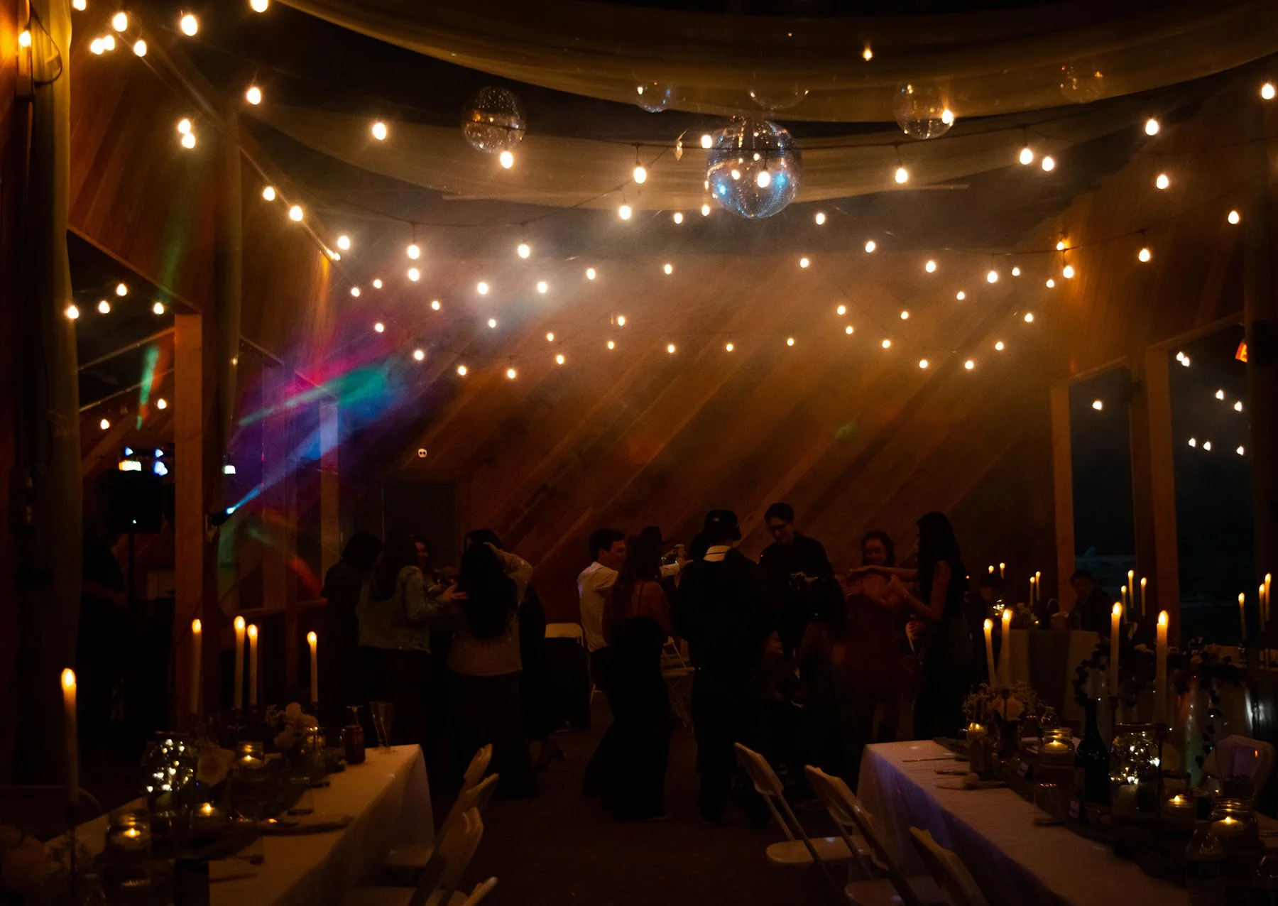 People dancing under string lights and disco balls in a decorated indoor party space with tables and candles.