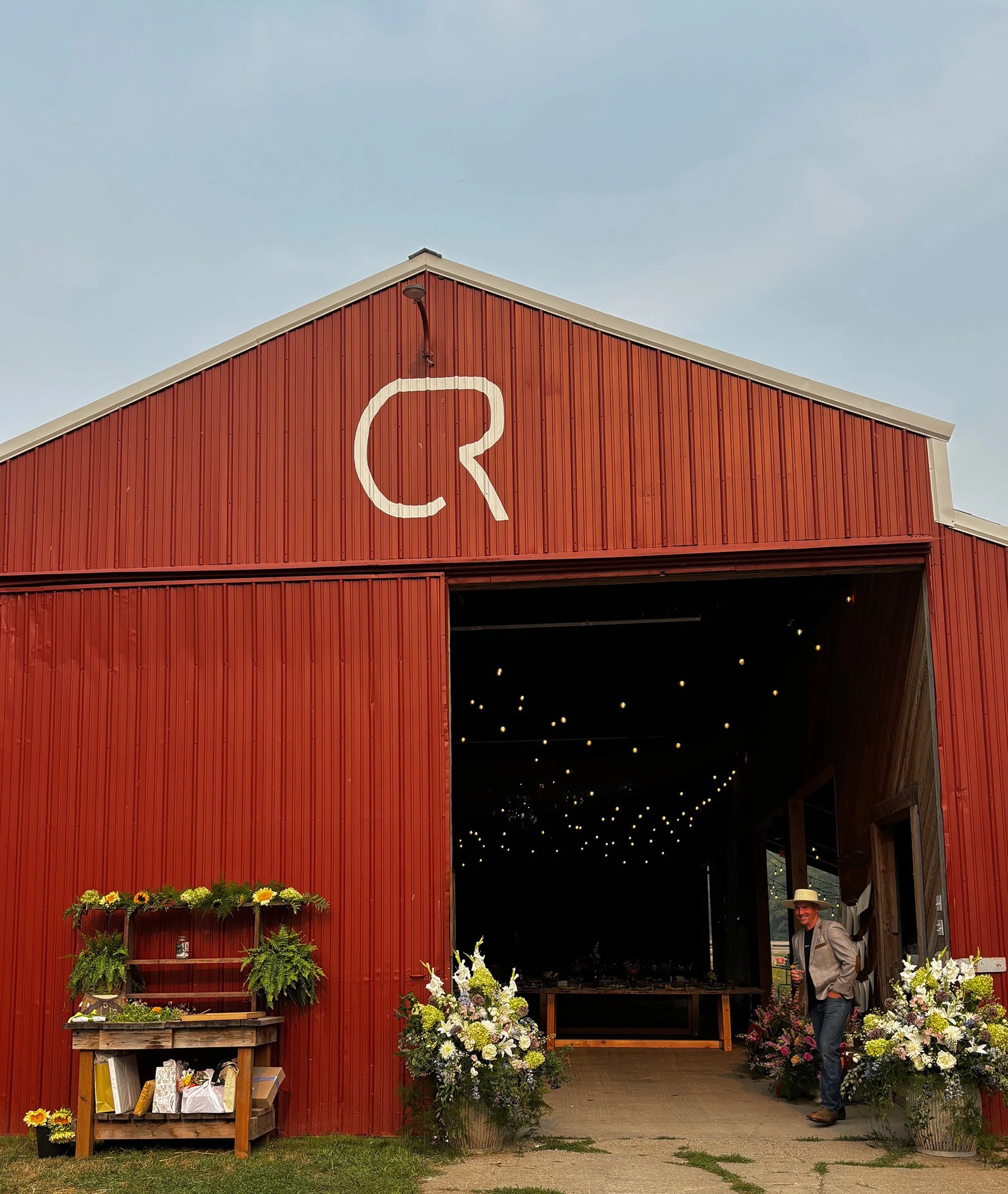 A red barn with an open entrance decorated with flowers, a man in a hat standing near the door, and string lights hanging inside.